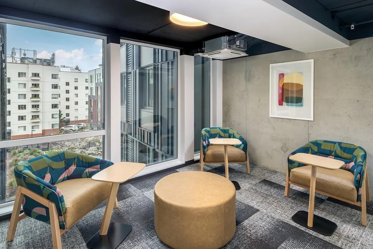 A modern lounge area featuring two colorful chairs with side tables and a circular coffee table. Large windows provide natural light, offering a view of an apartment building outdoors. Contemporary artwork hangs on the wall, complementing the minimalist design and concrete accents.