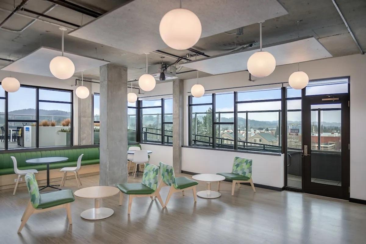 A modern indoor lounge area featuring several round white pendant lights hanging from the ceiling. The space includes green seating along a wall, small round tables, and white chairs with green accents. Large windows provide a view of the outdoors and let in natural light, creating a bright and open atmosphere.