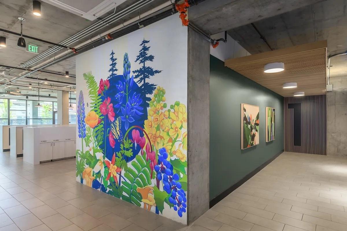 A vibrant mural featuring a mix of colorful flowers, plants, and trees, set against a white wall in a modern interior space. The surrounding area includes a green wall and framed art pieces, with industrial-style ceilings and lighting, enhancing the contemporary atmosphere of the environment.