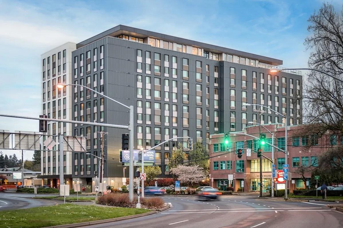 A modern, multi-story building at a street intersection with traffic lights, surrounded by greenery and other structures. The building features a contemporary design with large windows and varied facades, highlighting urban architecture. It's dusk, with soft lighting illuminating the exterior.