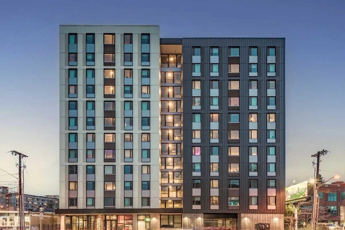 Modern multi-story building with a sleek design featuring large windows and balconies, illuminated by interior lights. The façade has a mix of light and dark panels, and the surrounding area includes urban elements like power lines and signage. The sky is transitioning to twilight.