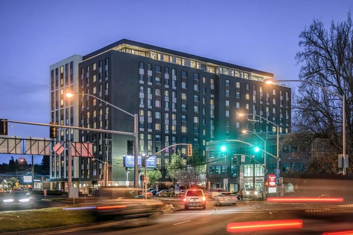 A modern multi-story building at dusk, with illuminated windows. Traffic lights and vehicles can be seen in the foreground, creating a dynamic urban scene. The building features a mix of dark panels and large glass windows, situated along a busy road with trees in the background.