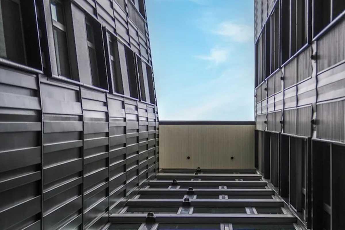 Looking up between two tall, modern buildings with metallic facades. The sky is visible at the top, creating a contrast with the darker structures on either side. The perspective emphasizes the height and architectural design of the buildings.