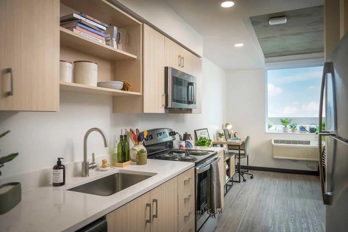 Modern kitchen with light wooden cabinets, a marble countertop, stainless steel appliances, and a window providing natural light. Shelves display books and decorative items. A workspace with a chair and desk is visible, along with green plants adding a fresh touch to the room.