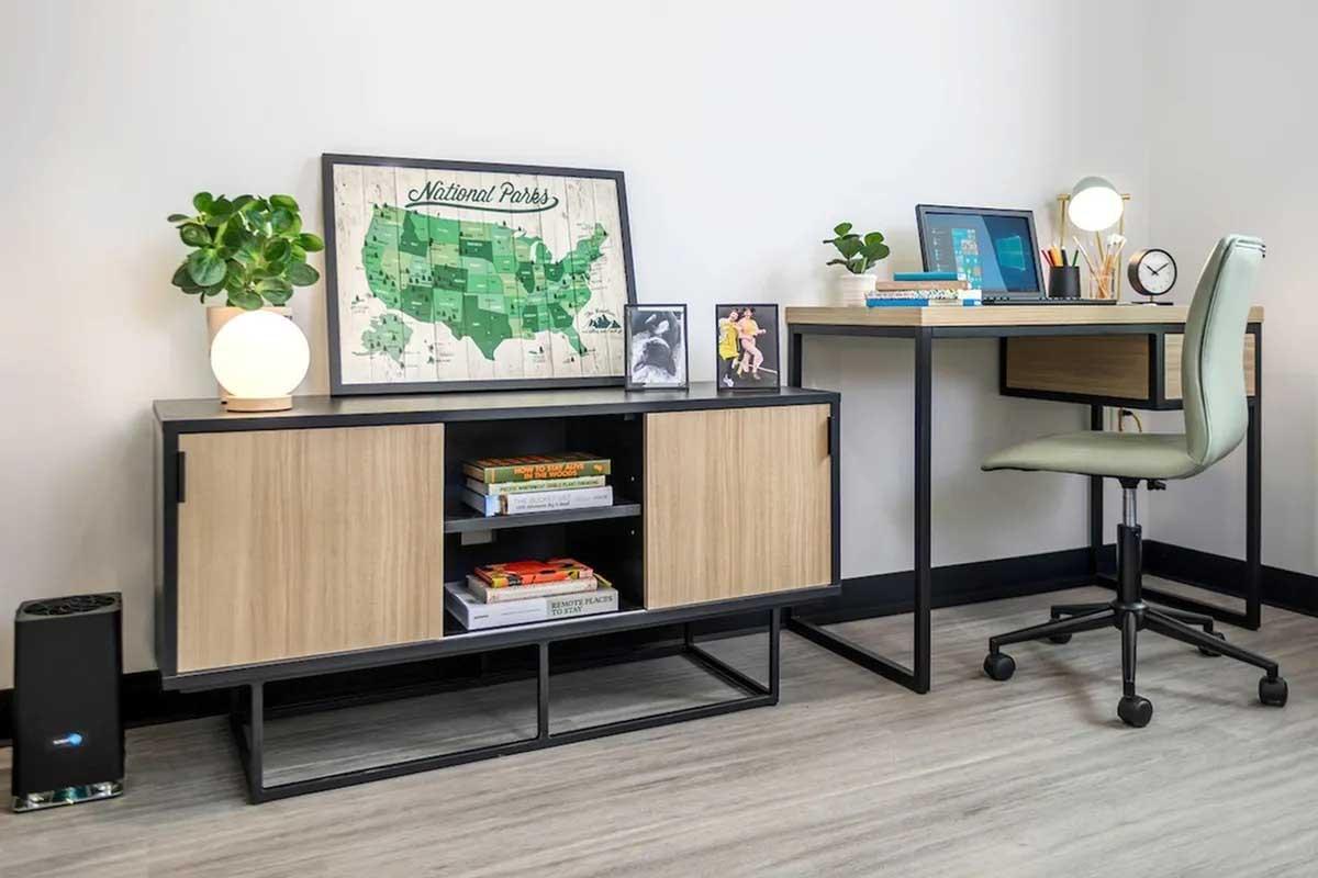 A modern workspace featuring a minimalist desk with a computer and stationery, a light green chair, and a light wood and metal storage unit. On the unit, there are books, a framed map of national parks, a photo frame, and two small plants. Soft lighting creates a cozy atmosphere in the room.