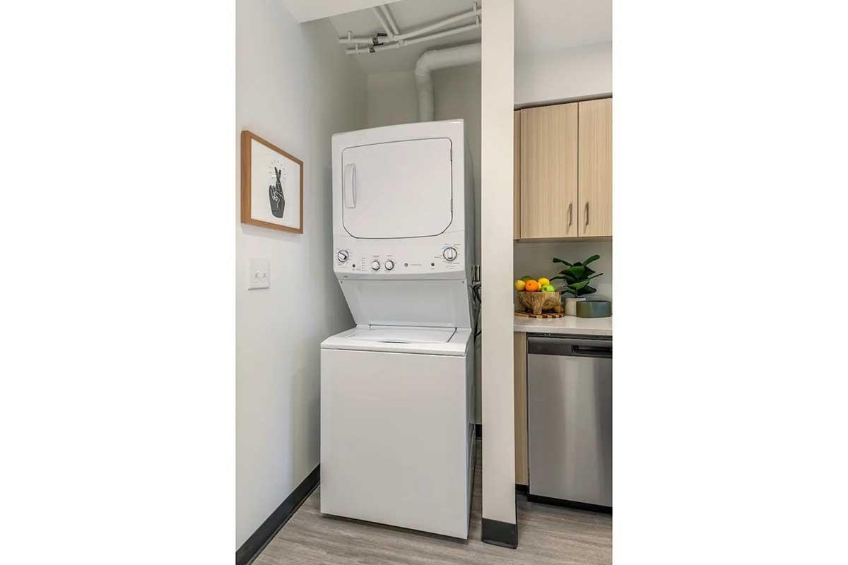 A stacked laundry unit consisting of a washer and dryer, positioned in a compact space between a wall and kitchen cabinetry, with a framed fruit artwork on the wall and a bowl of fruit visible on the countertop. The overall setting is modern and functional.