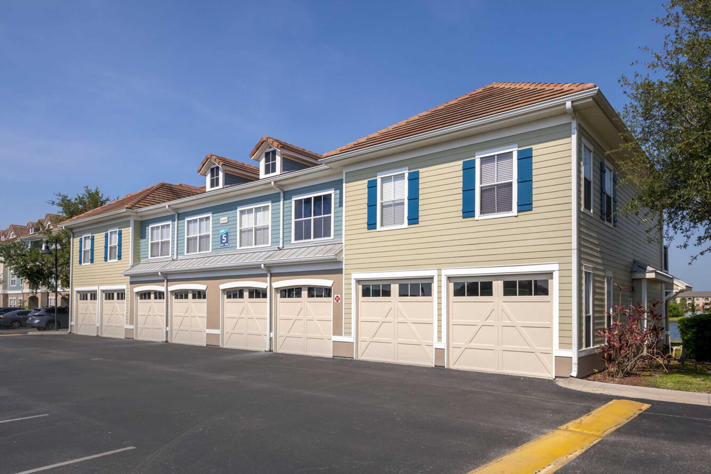 A row of two-story residential buildings with light-colored siding and blue accents. Each unit features a garage door at ground level, with several parking spaces in front. Clear blue sky overhead and greenery partially visible in the vicinity.