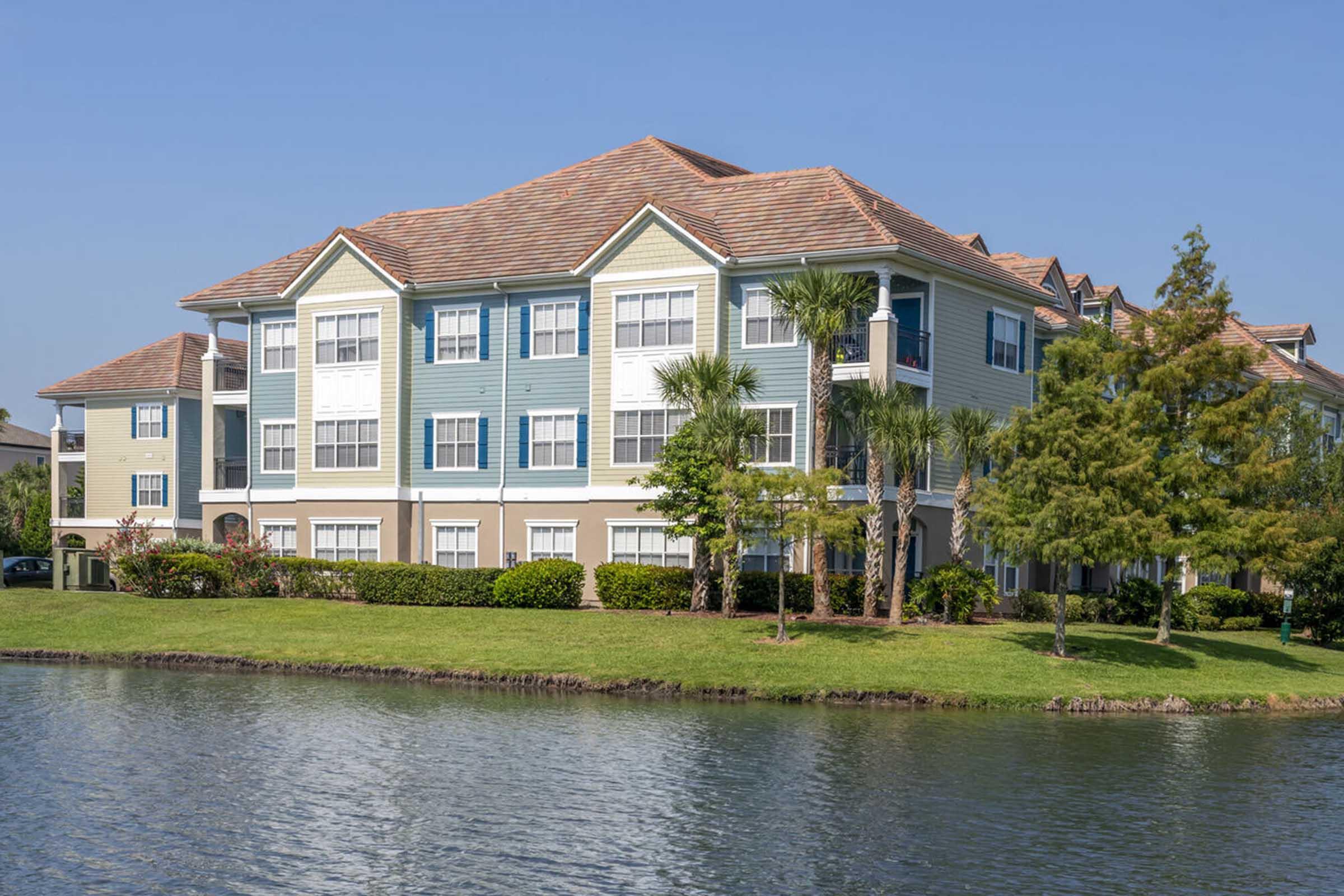 A multi-story residential building with a light blue and green facade, surrounded by lush greenery and palm trees. A calm body of water is in the foreground, reflecting the building and nearby landscaping under a clear blue sky.