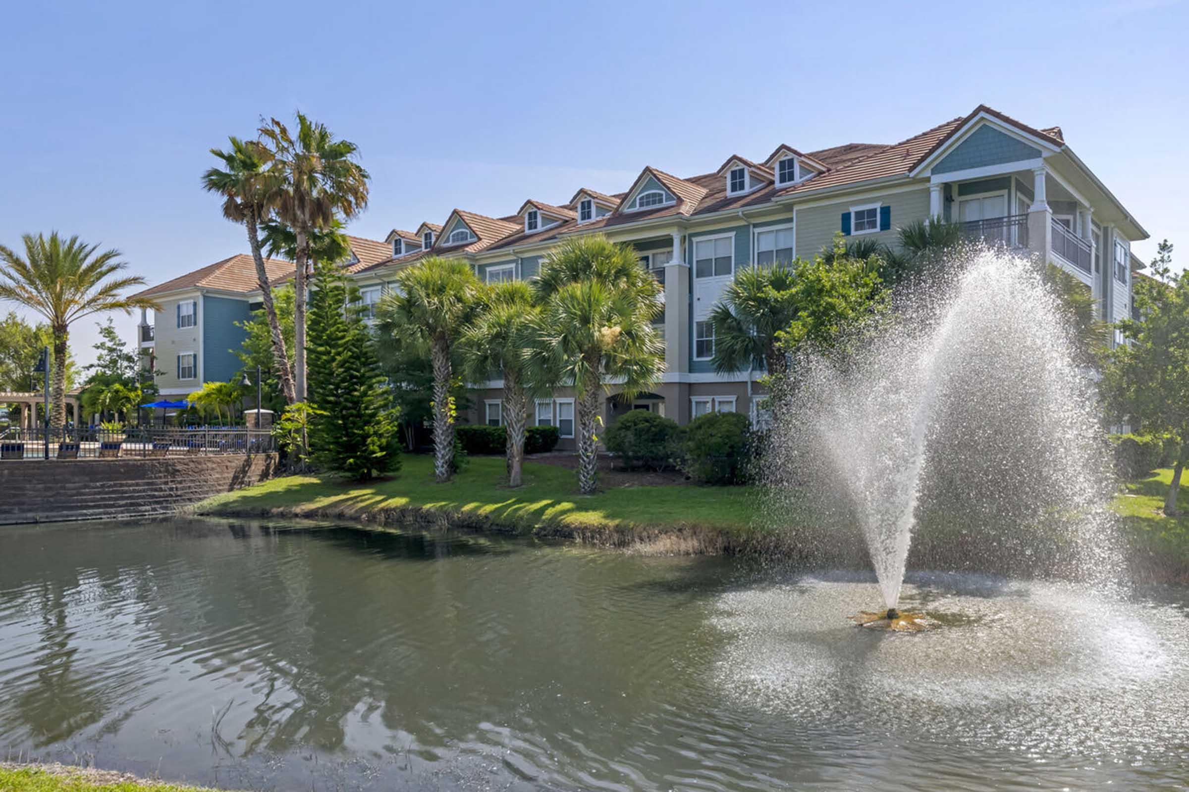 A tranquil scene featuring a green apartment building with multiple windows and a sloped roof, surrounded by palm trees. In the foreground, a pond with a fountain sprays water, creating a peaceful atmosphere. The clear blue sky adds to the serene setting.