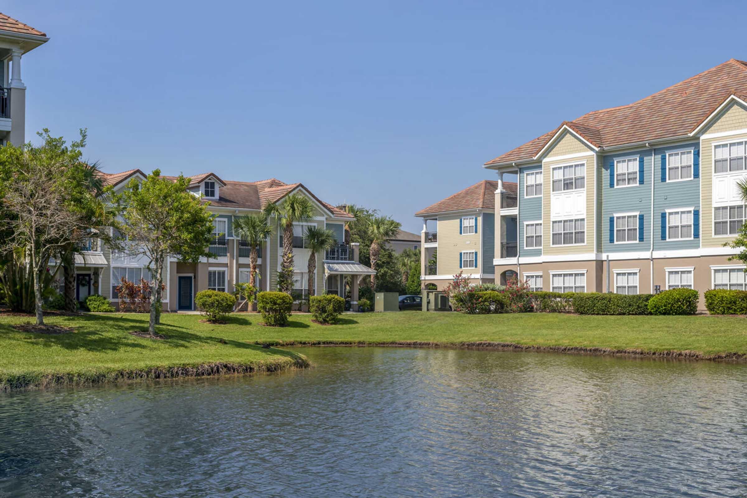 A serene view of an apartment complex beside a calm pond, surrounded by lush greenery. The building features a mix of traditional and modern architecture, with vibrant landscaping and palm trees, under a clear blue sky. The scene conveys a peaceful, suburban atmosphere.