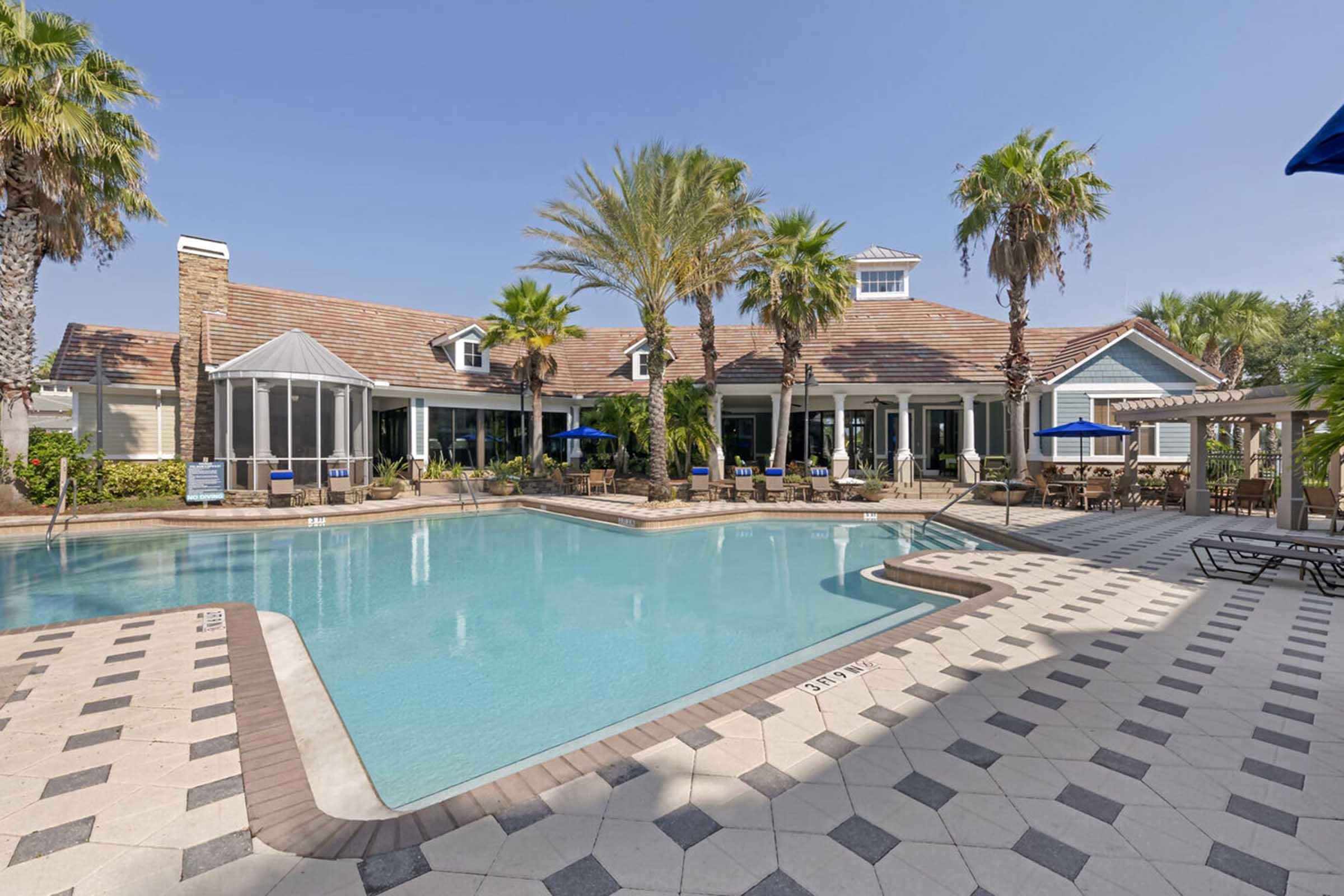 A serene pool area featuring a large swimming pool surrounded by palm trees and lounge chairs. The poolside has a decorative tile pattern, with a clubhouse in the background that includes large windows and shaded seating areas, all set against a clear blue sky.