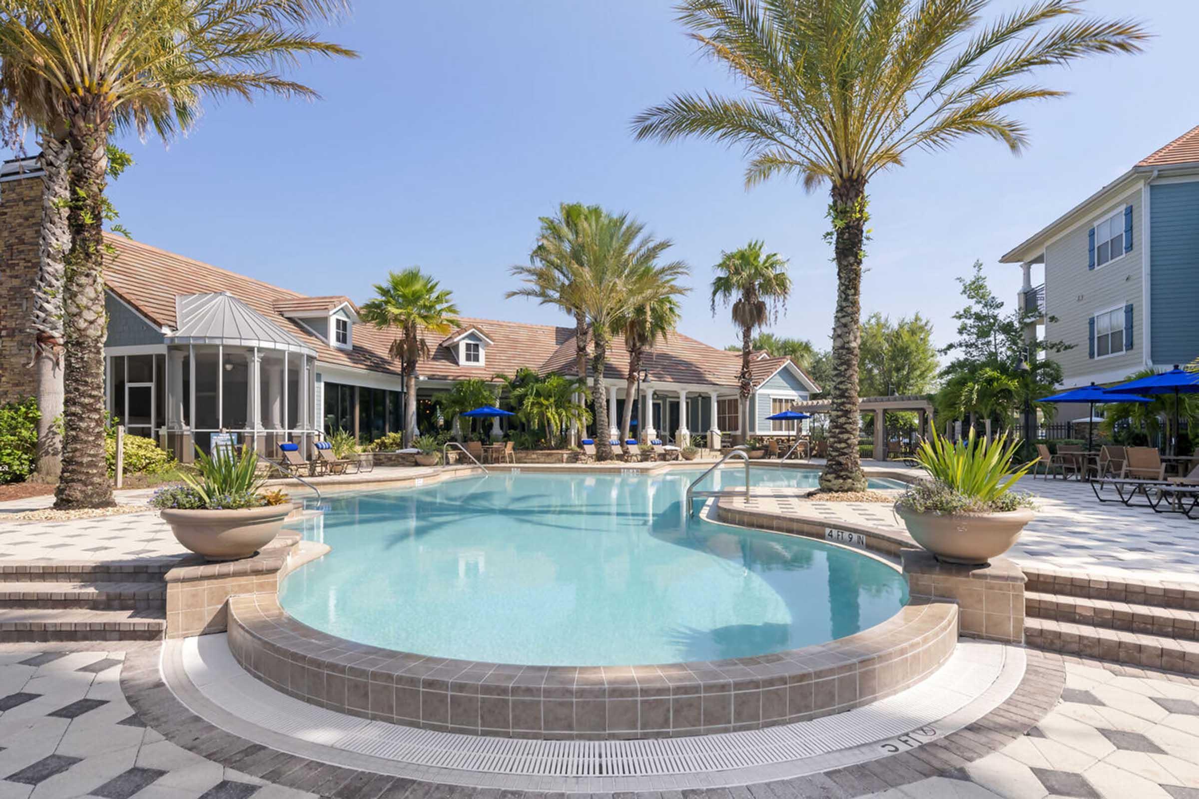 A resort-style swimming pool surrounded by palm trees and lounge chairs. Sunlight reflects off the clear water, and a few umbrellas provide shade. In the background, there are several buildings with a tropical design, creating a relaxing getaway atmosphere.