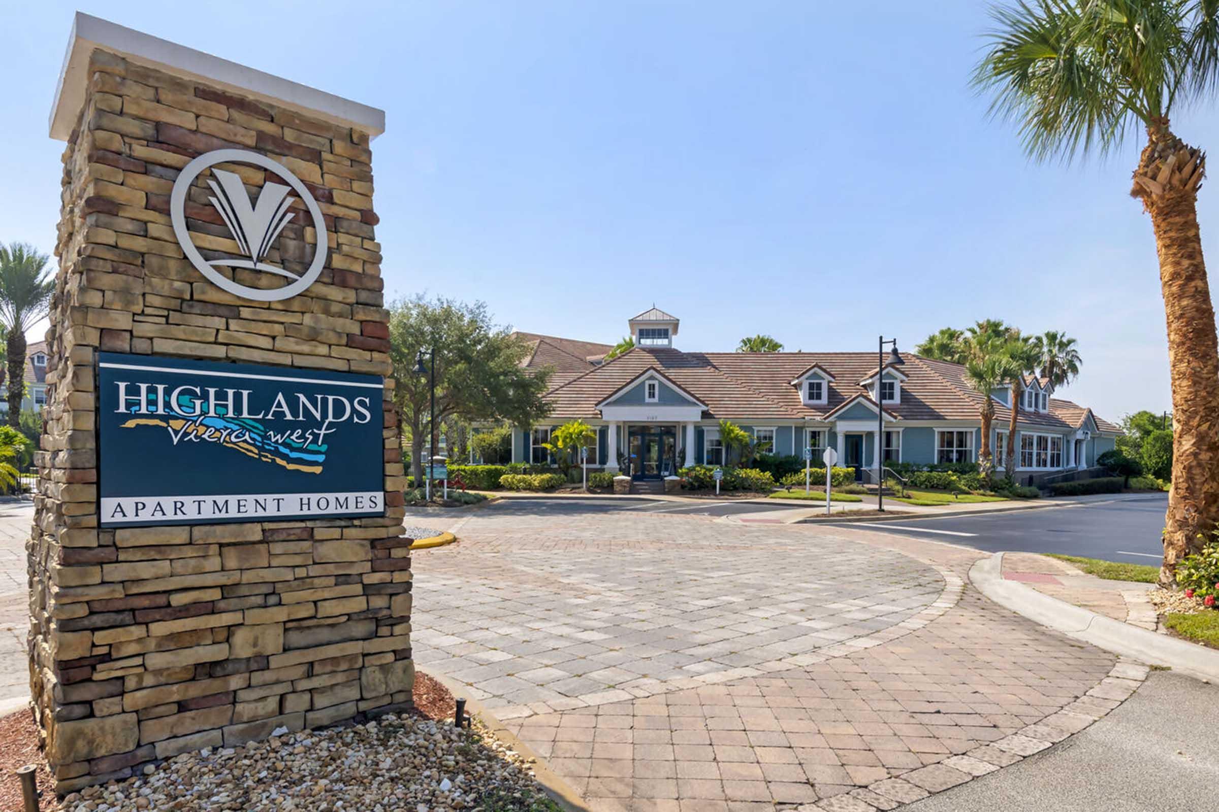 Sign for Highlands Vivere West apartment homes, featuring a stone structure with a modern logo. The scene includes lush palm trees and a well-maintained entrance with a paved road and landscaped details, creating a welcoming atmosphere. Clear blue sky overhead.