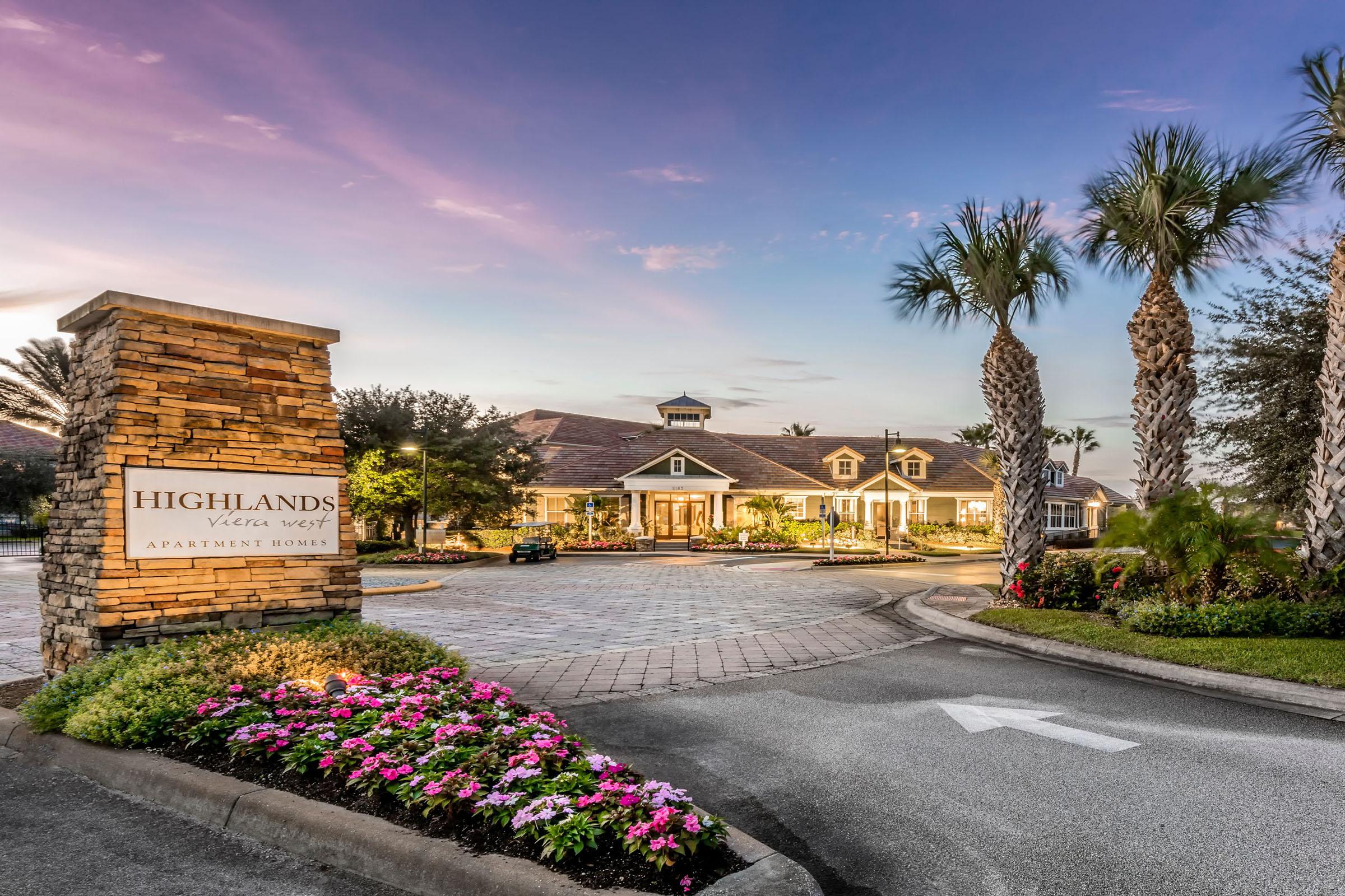A welcoming entrance to the Highlands Vista West apartment homes, featuring a stone sign surrounded by vibrant flowers, palm trees, and a well-maintained driveway leading to a beautifully designed building under a colorful sunset sky.