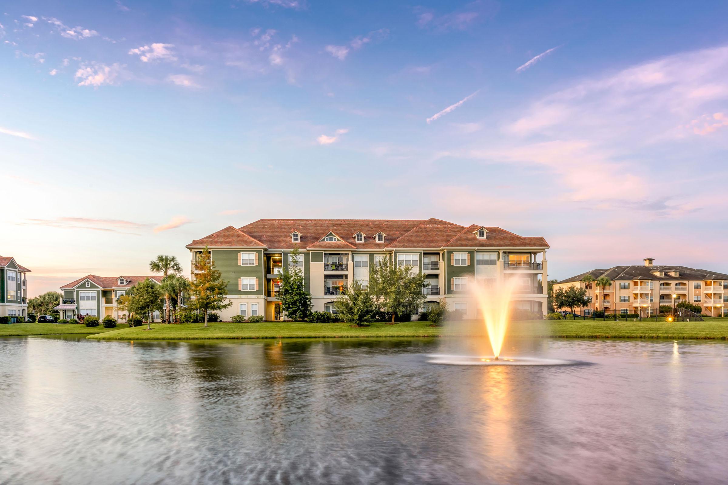 A serene lake scene featuring a fountain spraying water in the foreground. Behind the fountain, there are two multi-story apartment buildings with balconies, surrounded by lush greenery. The sky is painted with soft pastel hues of orange and blue as the sun sets, creating a tranquil atmosphere.