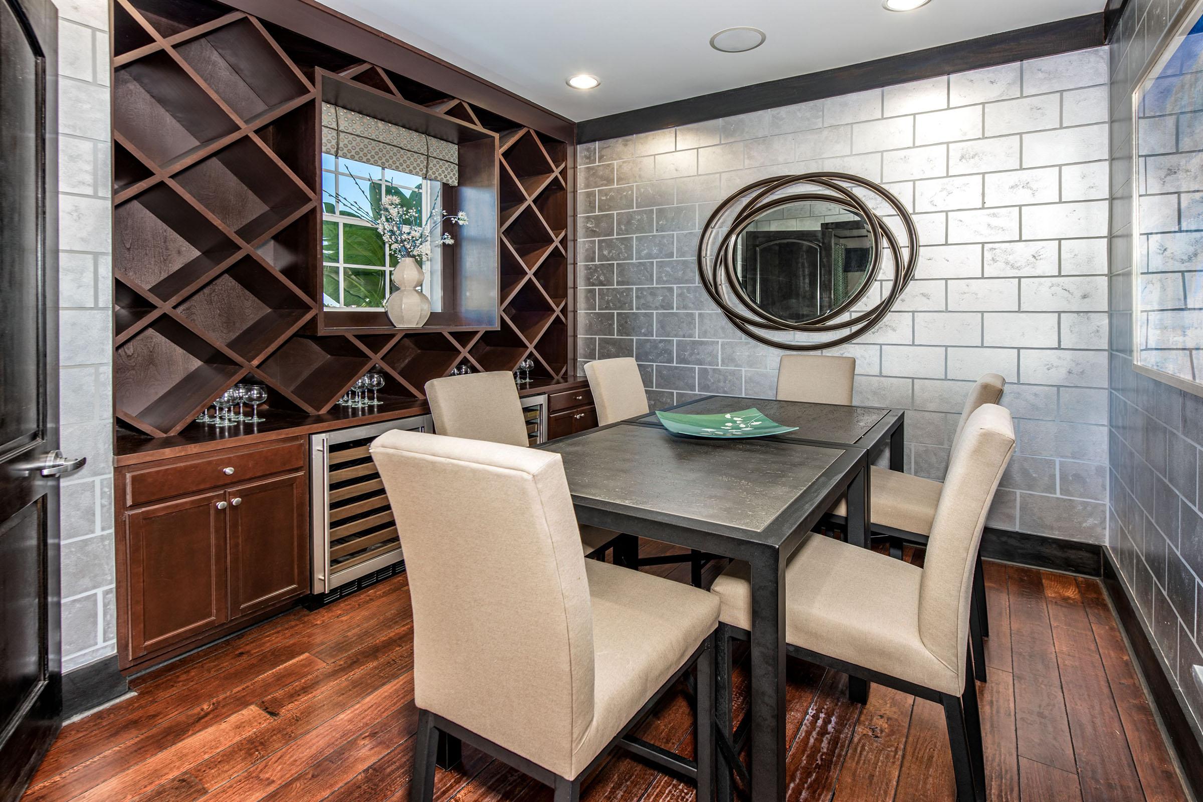 A stylish dining area featuring a dark wooden table surrounded by six beige chairs. Along one wall, there is a sleek wine rack and storage cabinets, with a large round mirror and modern decor. The walls are a mix of gray tiles and smooth finishes, complemented by dark hardwood flooring and natural light from a window.