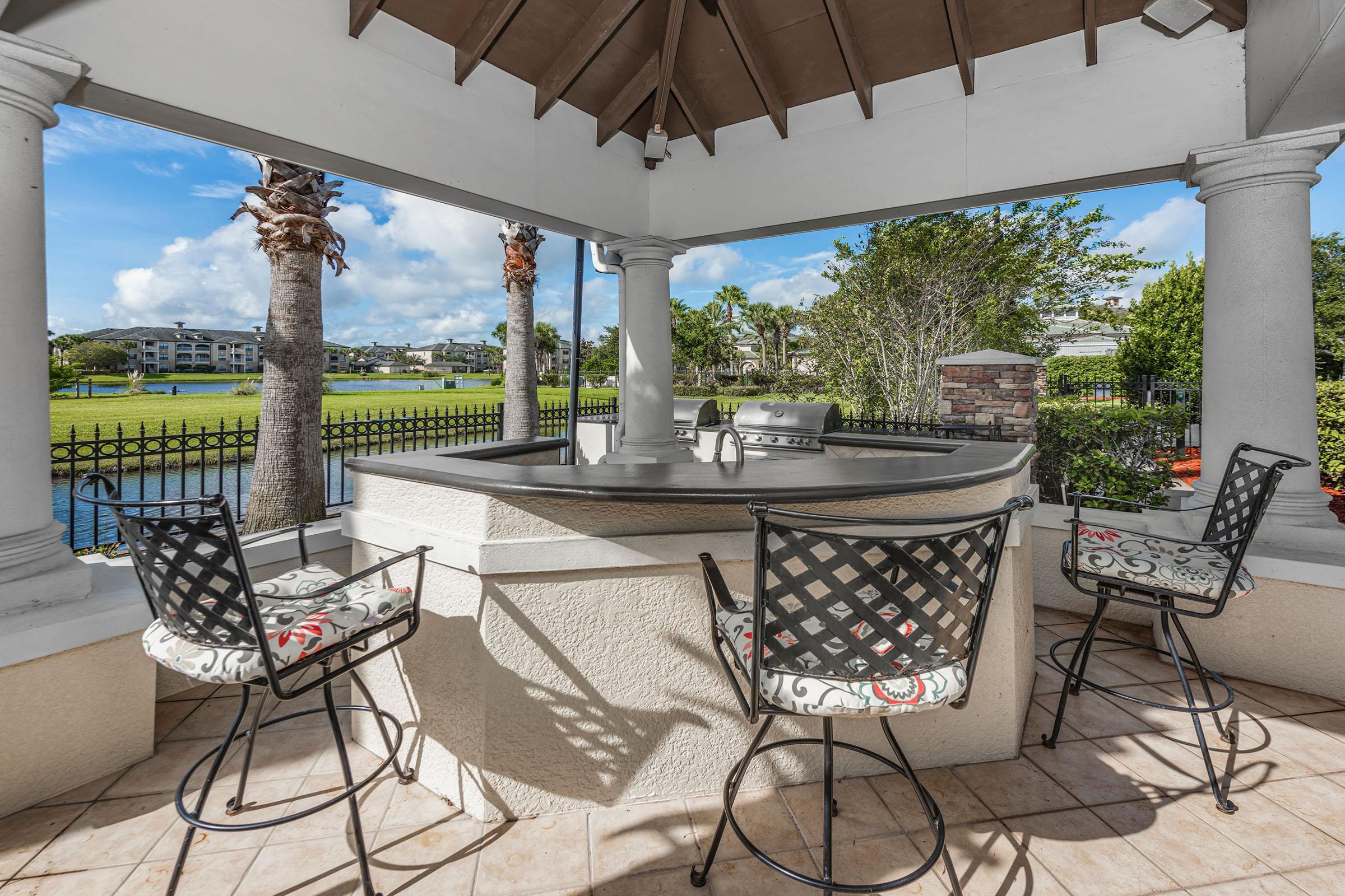 A shaded outdoor bar area featuring a curved countertop with a grill, surrounded by four decorative high-back chairs. In the background, a serene view of a lake and palm trees, with blue skies and fluffy clouds creating a relaxing atmosphere.