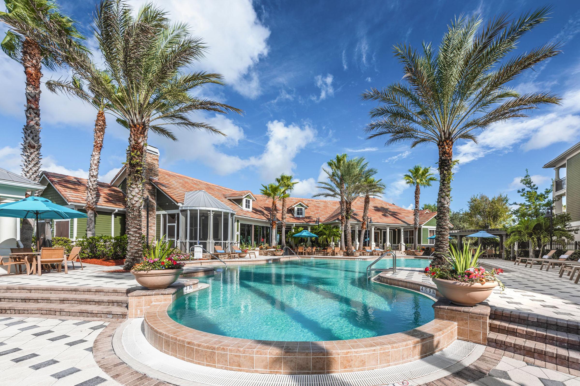 A beautifully designed pool area surrounded by palm trees and lounge chairs. The clear blue water reflects the sky, and there are green umbrellas providing shade. In the background, a modern building complements the tropical setting, making it an inviting space for relaxation and socializing.