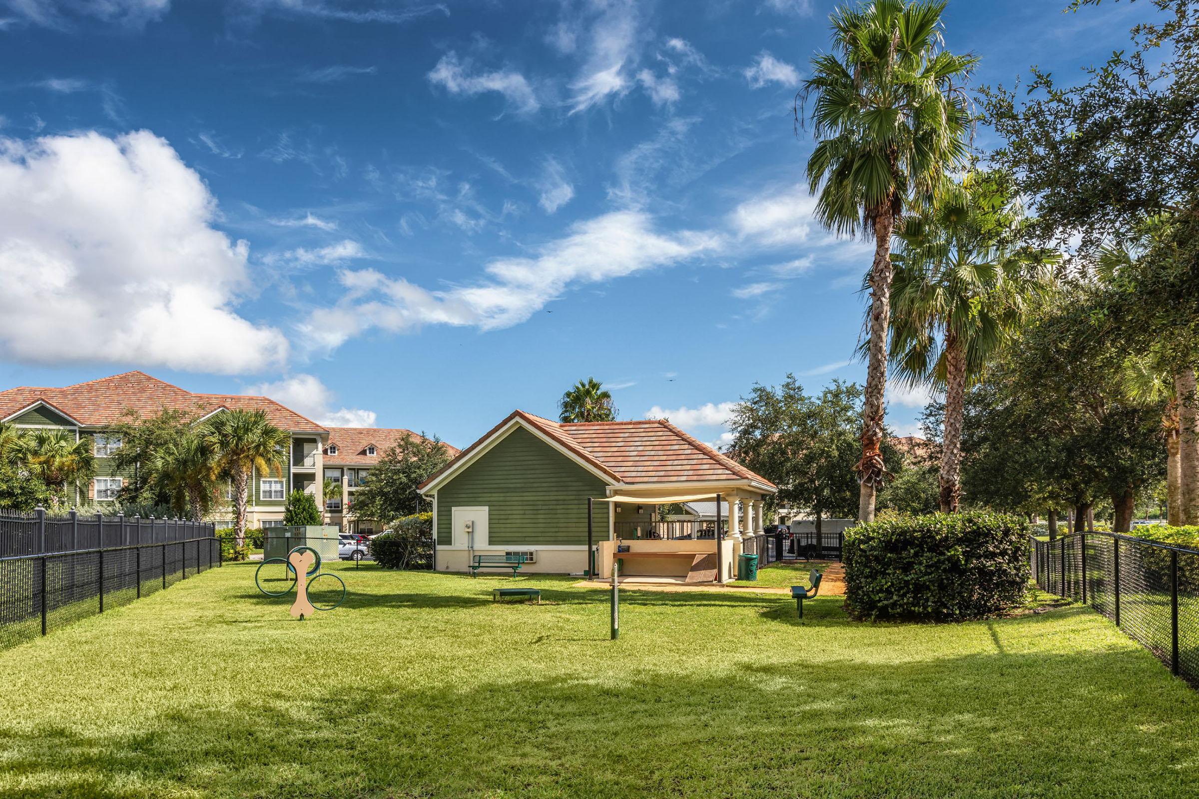A grassy area with palm trees and a small green building, possibly a community center. There are fitness stations visible, and nearby residential buildings in the background. The sky is blue with fluffy clouds, creating a sunny and inviting atmosphere.