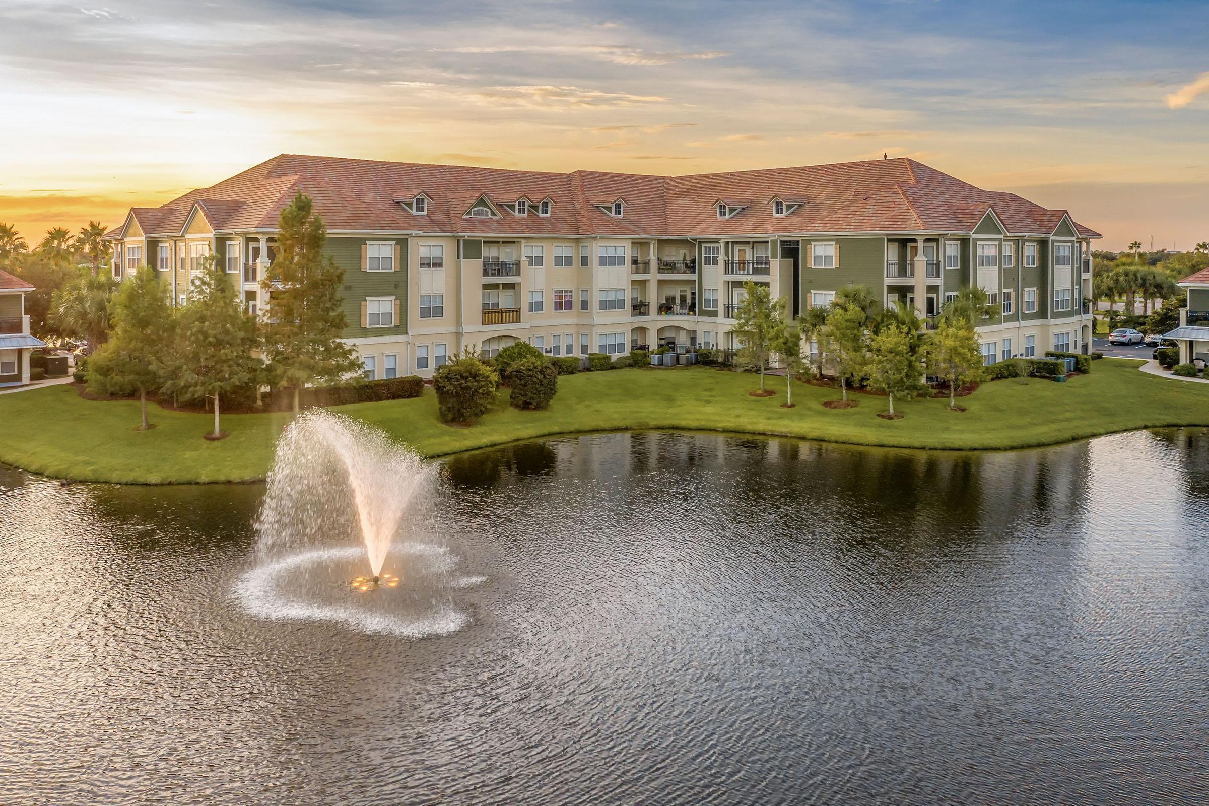 A scenic view of a multi-story apartment building surrounded by lush greenery, situated next to a calm pond with a decorative fountain. The image captures the sunset, casting a warm glow over the scene, enhancing the tranquil atmosphere of the residential area.
