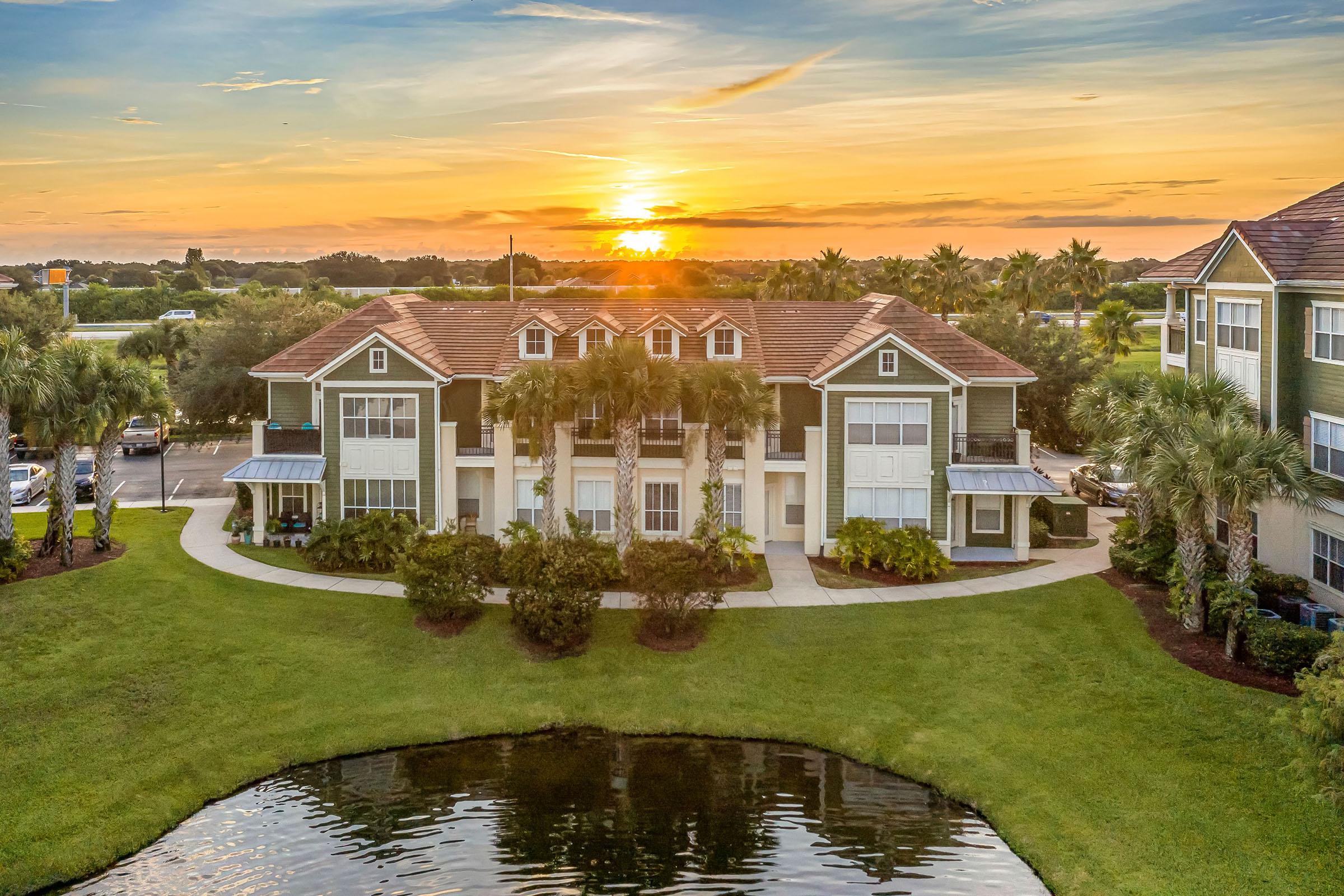 A scenic view of a well-maintained residential building surrounded by a green lawn and palm trees, reflecting in a small pond. The sun sets in the background, casting a warm glow over the landscape, with a clear sky and scattered clouds.