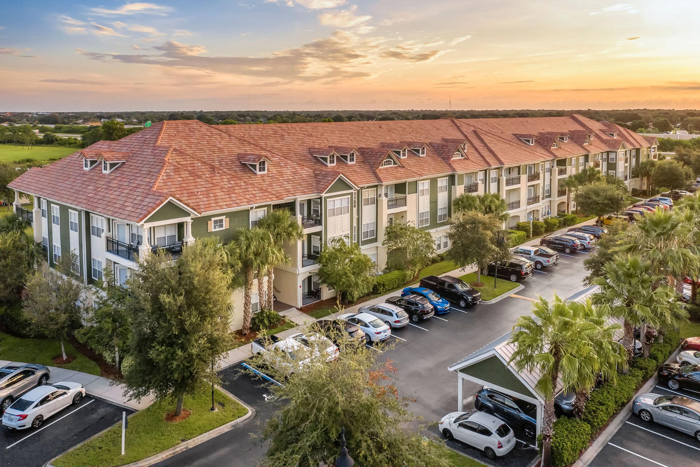 A multi-story residential building with a reddish-brown roof, surrounded by palm trees and a parking lot filled with cars. The scene is set during sunset, with a colorful sky in the background, creating a warm and inviting atmosphere.
