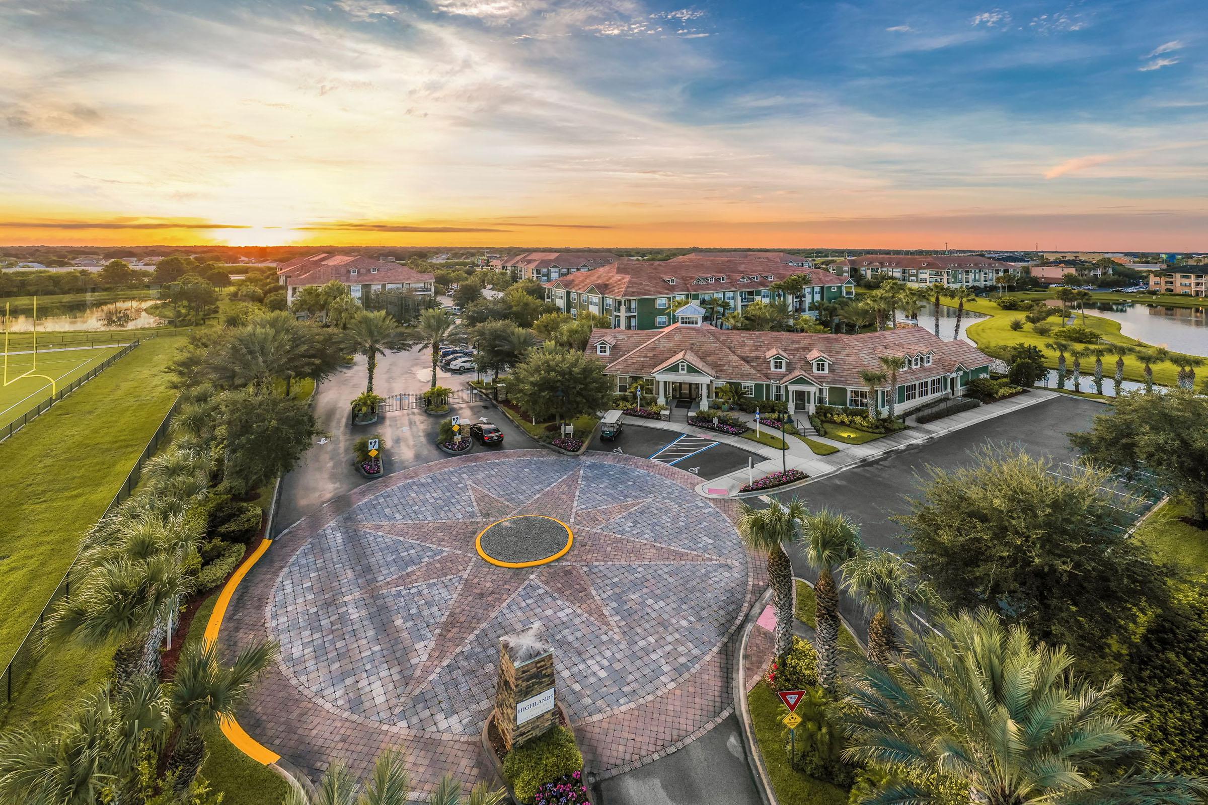 Aerial view of a landscaped residential community at sunset. The scene features a circular driveway with a star-shaped design, palm trees lining the entrance, and buildings in the background. A lake is visible, reflecting the colorful sky, creating a serene atmosphere.