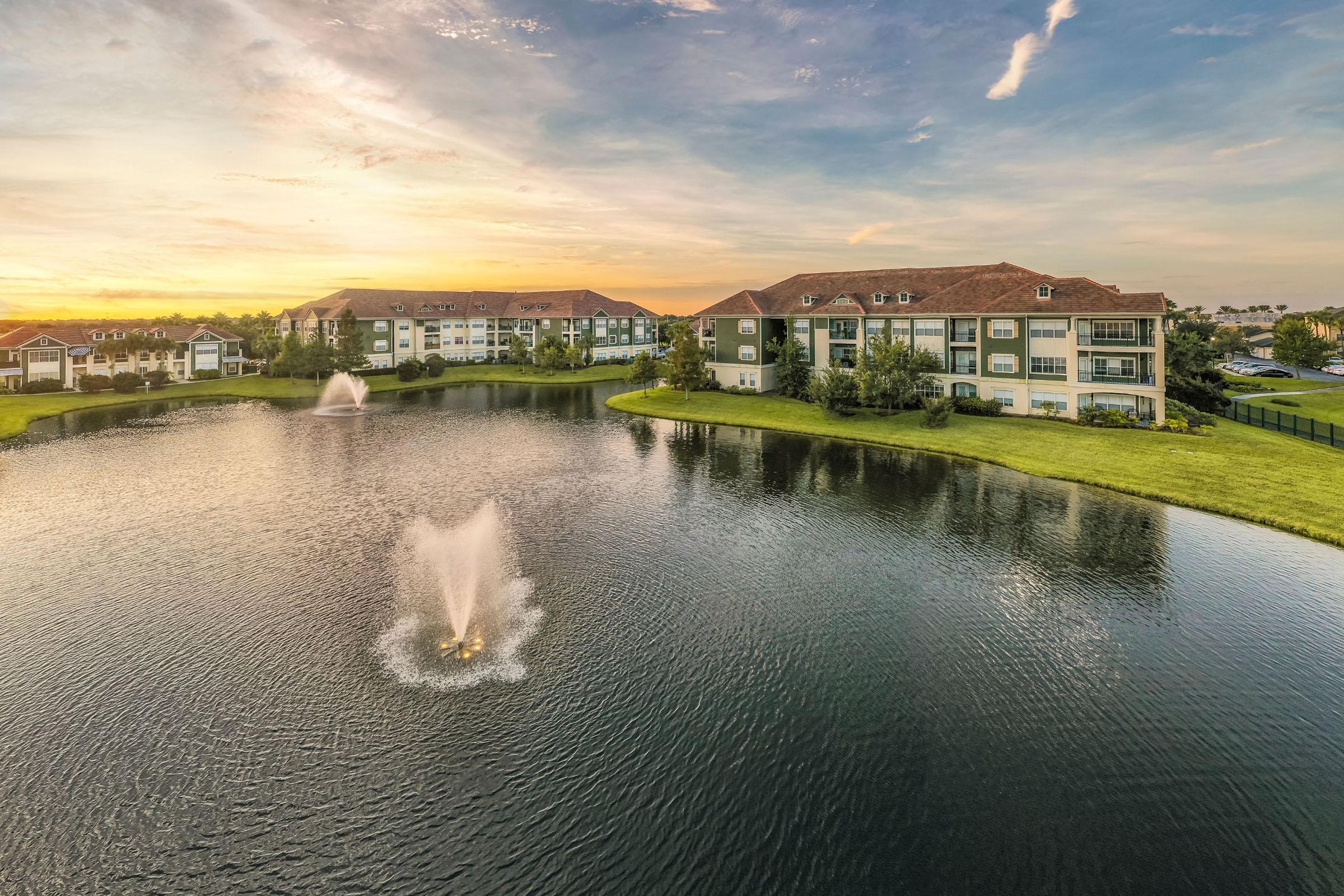 A scenic view of a tranquil lake with a fountain in the foreground, surrounded by lush greenery and modern apartment buildings. The sky displays vibrant colors of a sunset, creating a peaceful atmosphere reflected in the water. Ideal for showcasing a serene living environment.
