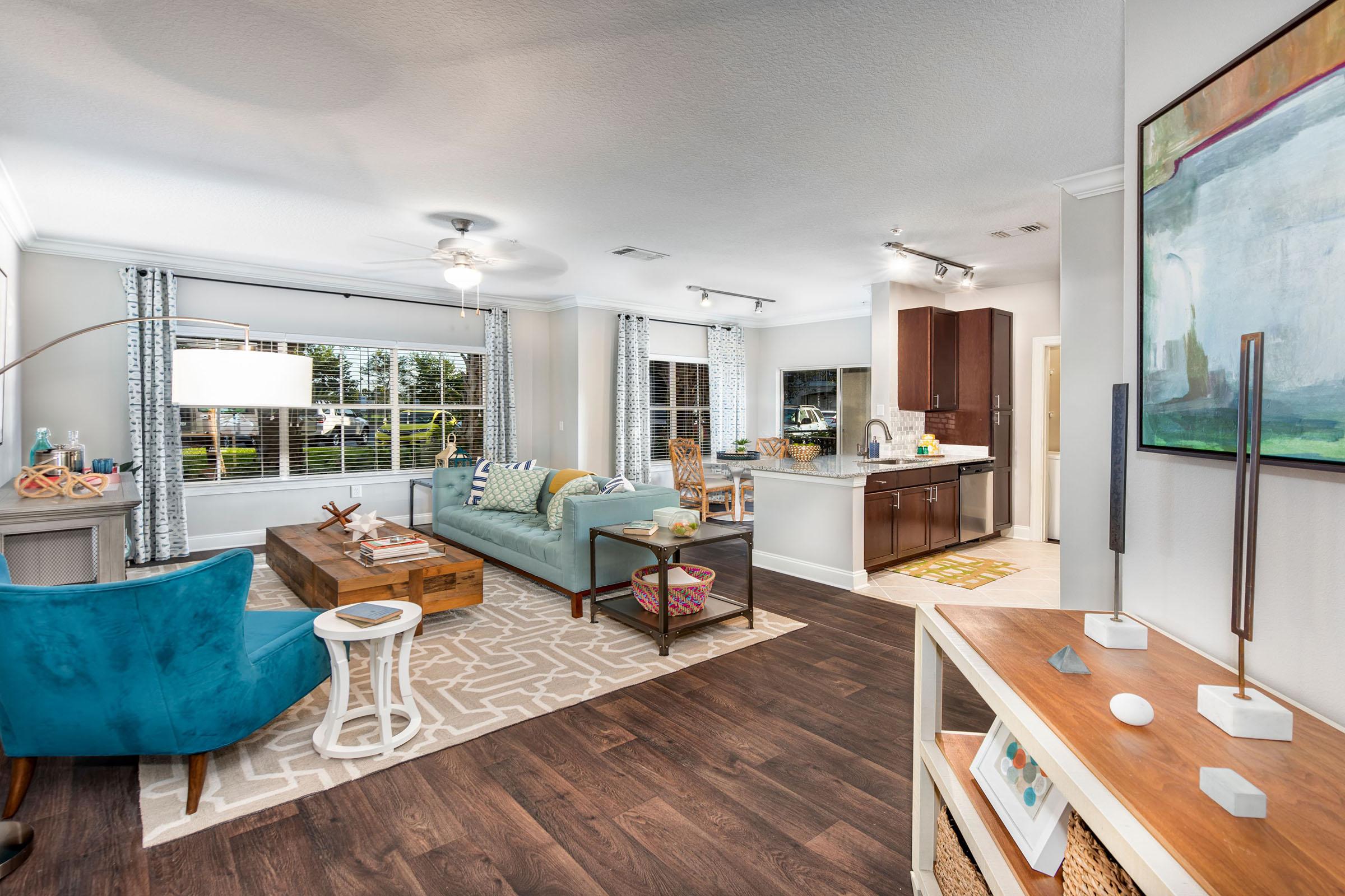 A modern living room featuring a light blue sofa, wooden coffee table, and an accent chair. The space has large windows with natural light, decorative items on shelves, and a tasteful rug. The kitchen is visible in the background with dark cabinetry and light-colored countertops.