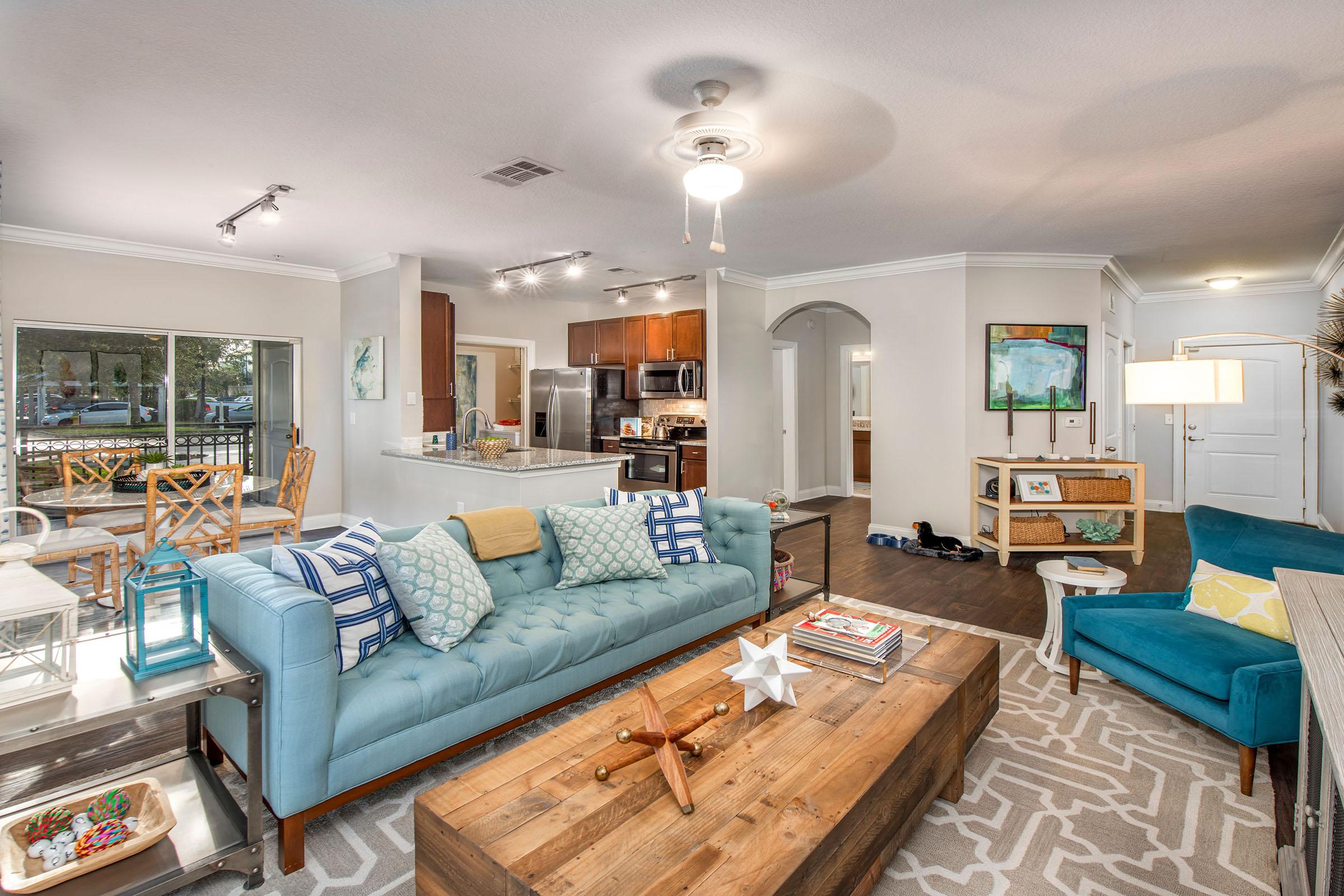 A cozy living room featuring a light blue sofa with colorful cushions, a wooden coffee table, and a teal accent chair. The space includes a dining area with stylish chairs and a modern kitchen visible in the background. Natural light shines through a window, enhancing the warm and inviting atmosphere.