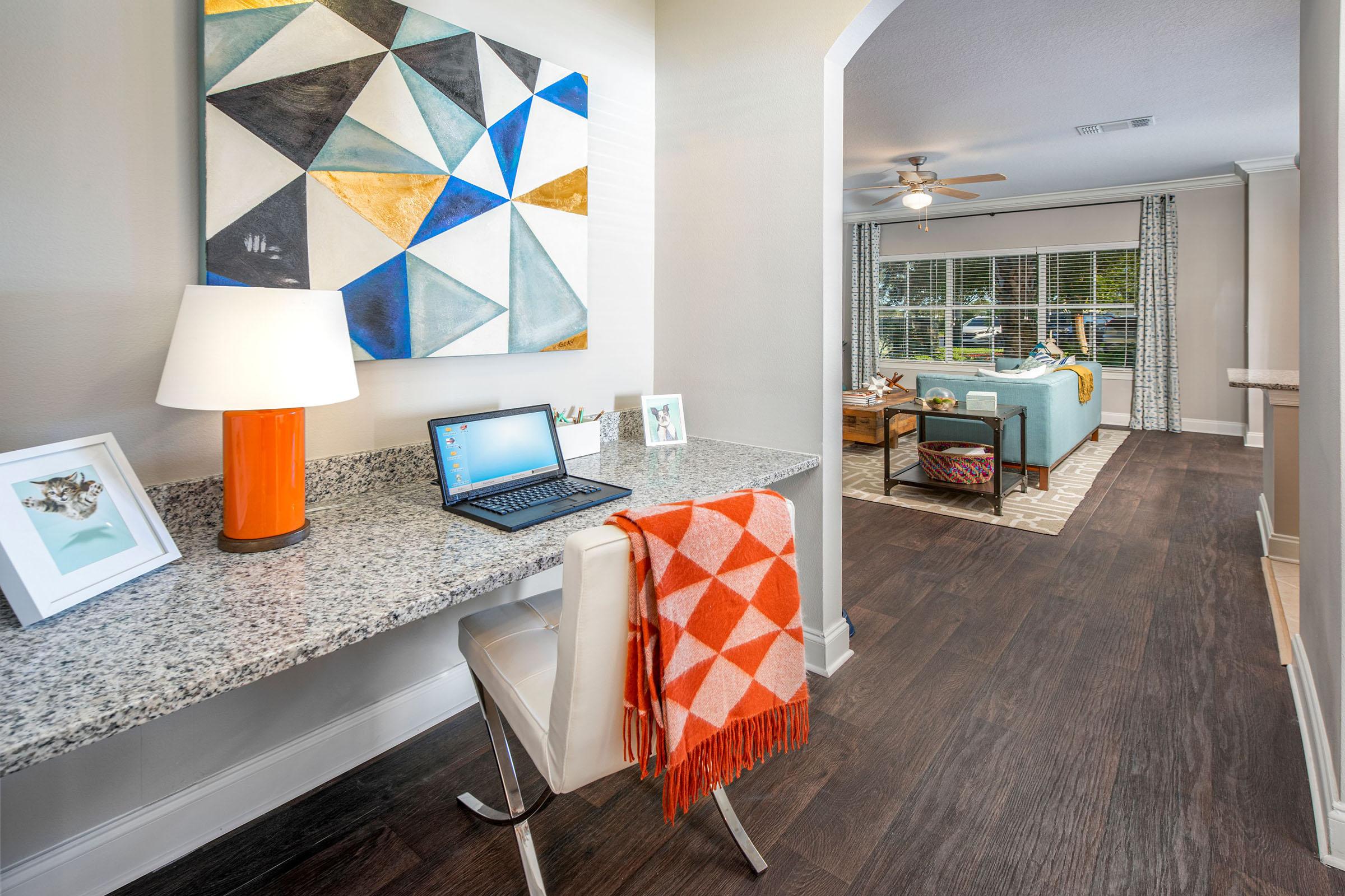 A modern home interior featuring a small workspace with a granite-topped desk, an orange lamp, and a laptop. A cozy throw blanket with geometric patterns is draped over the chair. In the background, a living area is visible with a light blue couch, a coffee table, and large windows letting in natural light.