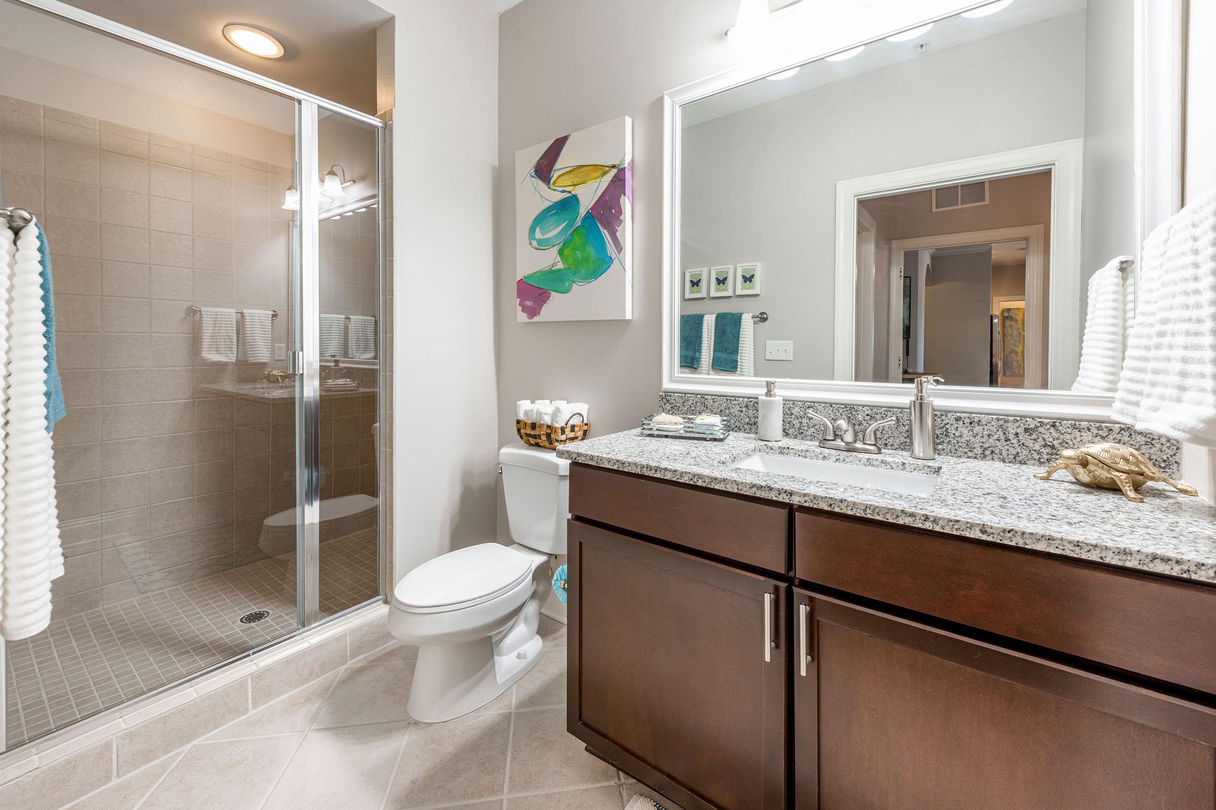 A modern bathroom featuring a glass-enclosed shower, a white toilet, and a dark wood vanity with a granite countertop. There is a large mirror above the sink, decorative wall art, and neatly arranged towels. The floor is tiled in a neutral color, creating a clean and stylish look.
