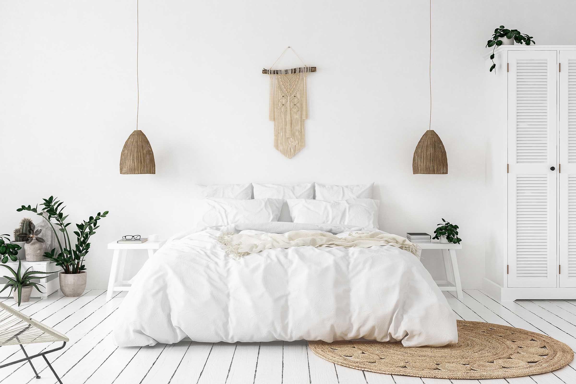 A cozy, minimalist bedroom featuring a large white bed with fluffy bedding, wooden accents, and two hanging light fixtures. There's a round woven rug on the floor, green plants in the corners, and a tall white cabinet with louvered doors, creating a serene and inviting atmosphere.