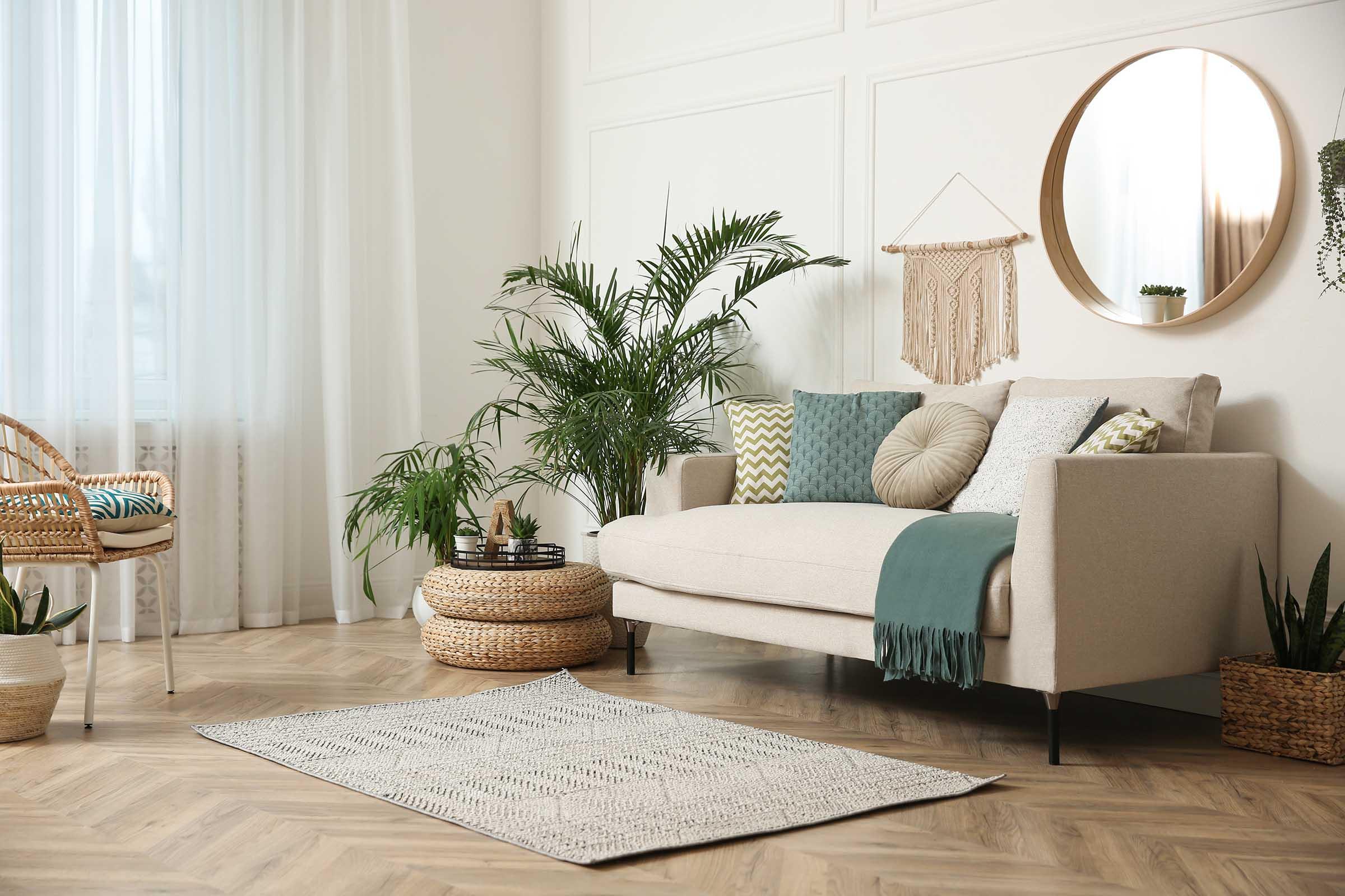 Cozy living room featuring a light-colored sofa with decorative pillows, a round mirror on the wall, and various indoor plants. A woven rug lies on the wooden floor, complemented by a rattan chair. Natural light pours in through sheer curtains, creating a warm and inviting atmosphere.