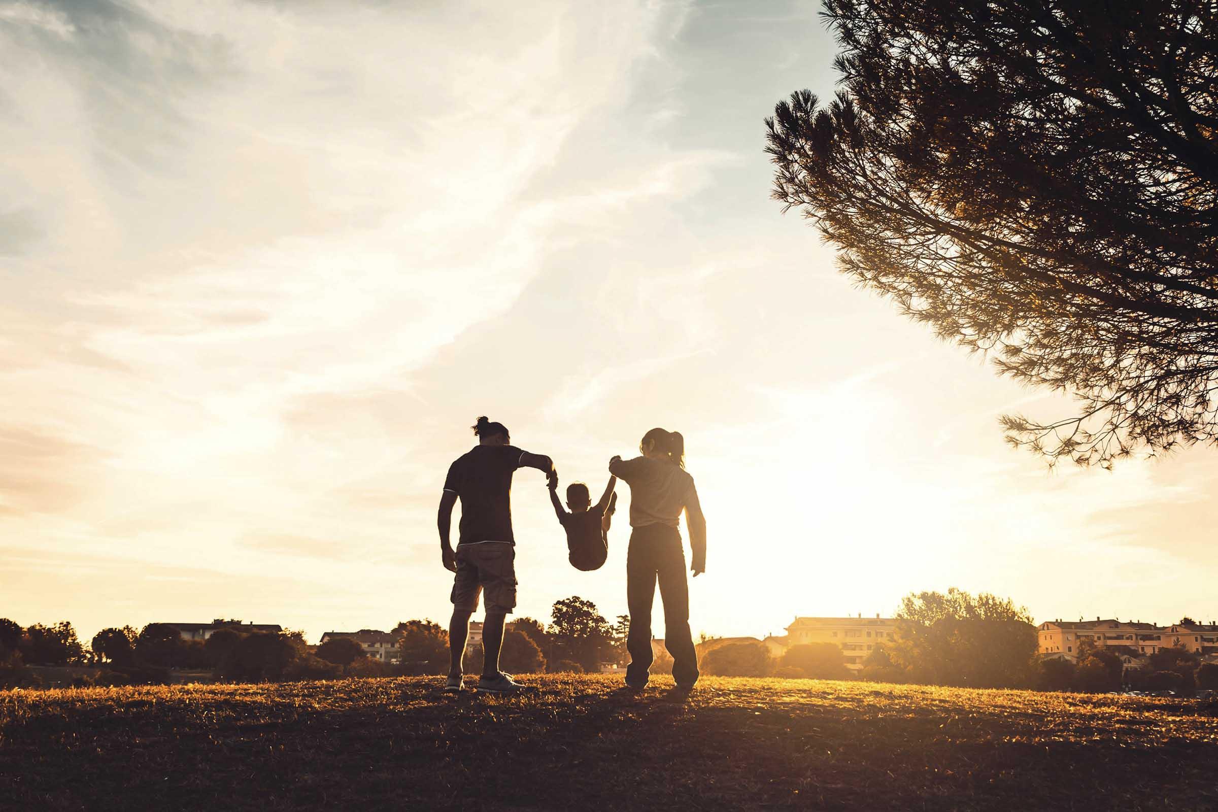A silhouette of a man and a woman holding hands while lifting a small child between them. They are standing on a grassy hill against a sunset background, with trees and buildings in the distance, creating a warm, joyful family scene.
