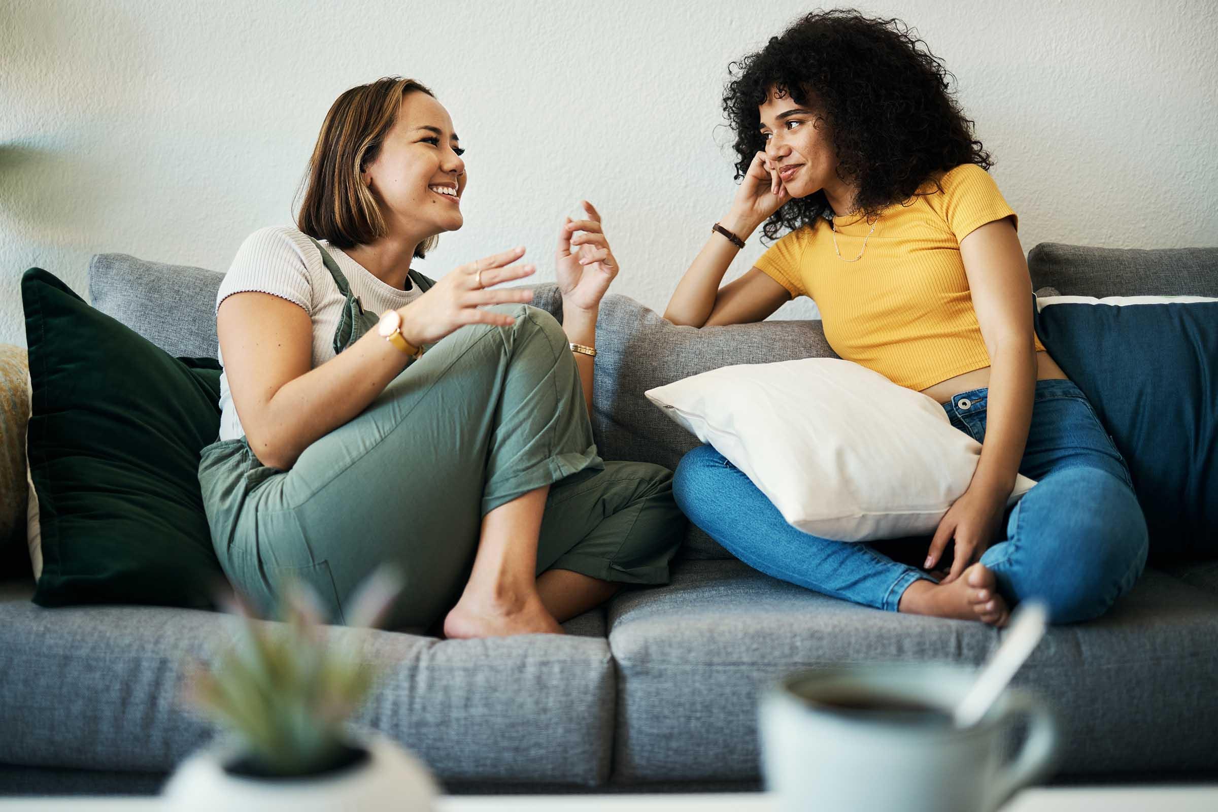 Two women are sitting on a gray sofa, engaged in a lively conversation. One woman, wearing a white top and green overalls, smiles and gestures while speaking. The other woman, in a yellow shirt and blue jeans, listens thoughtfully, resting her chin on her hand. There are decorative pillows and a coffee mug on a table nearby.