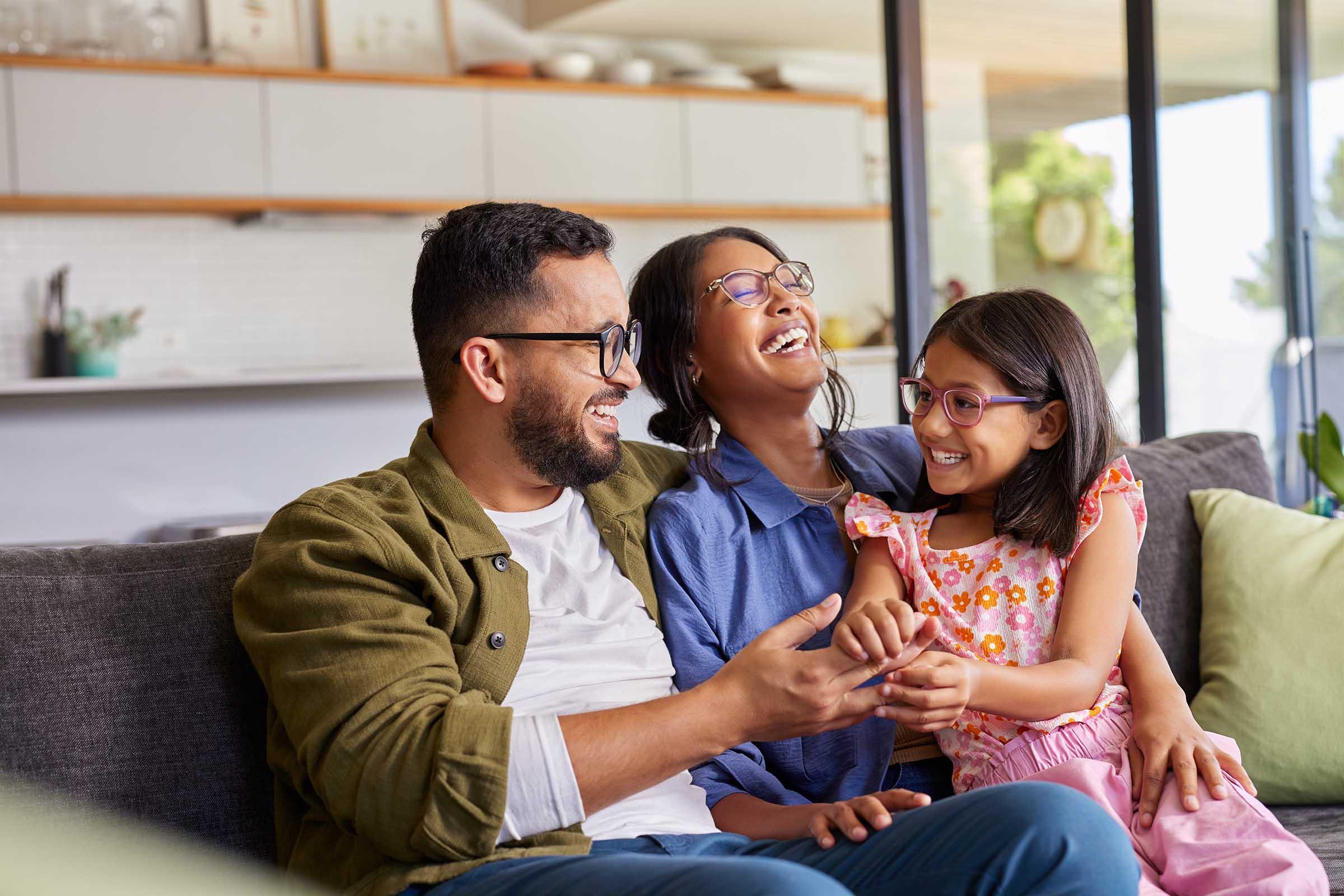 A joyful family sitting on a couch, laughing together. A man and a woman, both wearing glasses, are sharing a light-hearted moment with a young girl. The girl is smiling and holding the man's hand, while the woman looks at her with affection. The setting is a bright, modern living space.