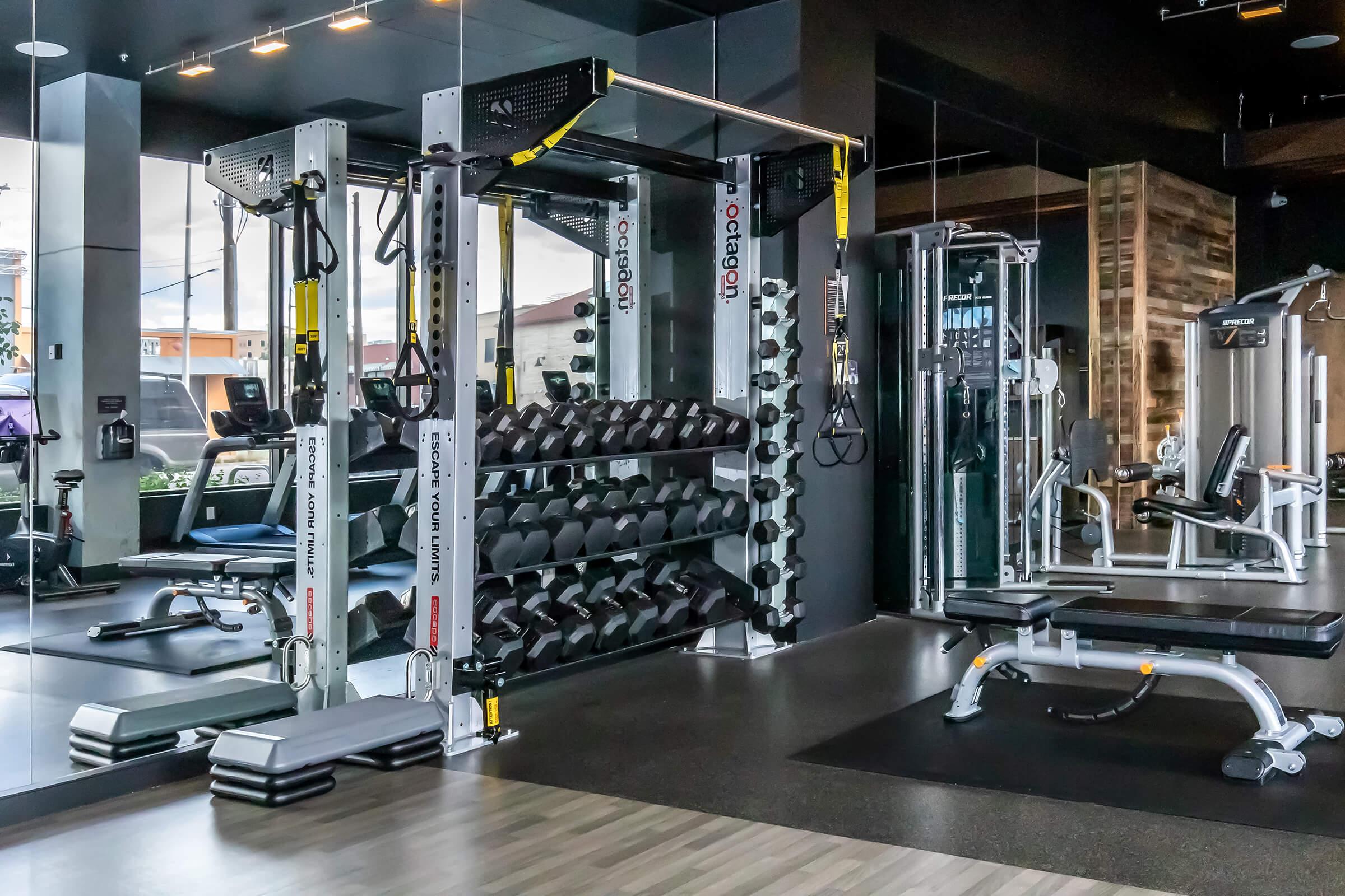 A modern gym interior featuring a variety of workout equipment, including a multi-use training machine, dumbbells, a weight bench, and cardio machines. Large mirrors reflect the space, and natural light enters through windows, creating a bright and inviting atmosphere for fitness enthusiasts.