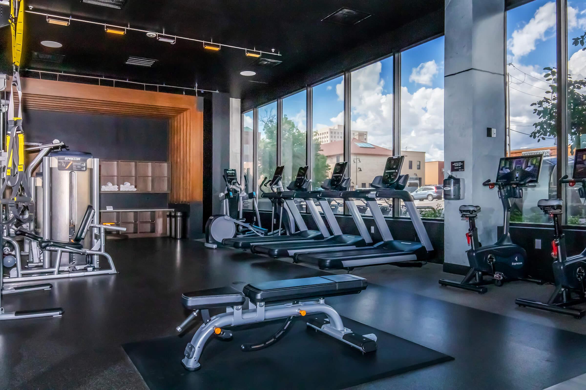 A modern gym interior featuring exercise equipment, including treadmills and stationary bikes, arranged near large windows that let in natural light. The floor is dark and sleek, and there are benches and weight machines visible, creating a spacious and inviting workout environment.