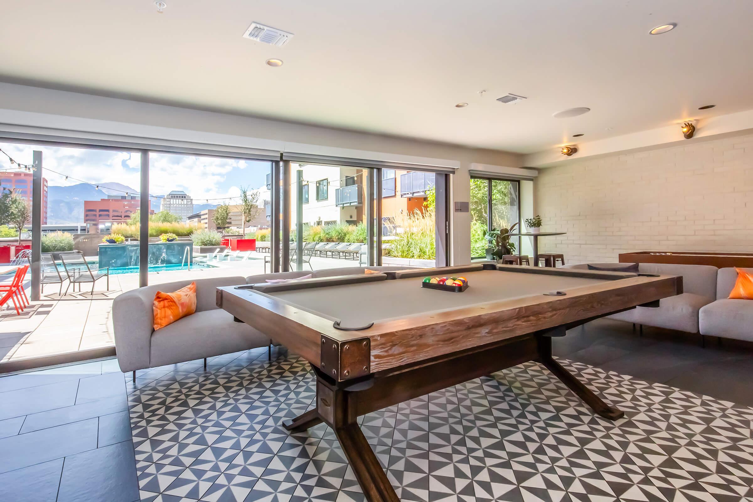 A modern indoor lounge area featuring a billiard table with colorful pool balls on top. Comfortable gray sofas with orange cushions are positioned nearby. Large windows offer a view of an outdoor pool surrounded by greenery and bright lounge chairs, providing a bright and inviting atmosphere.