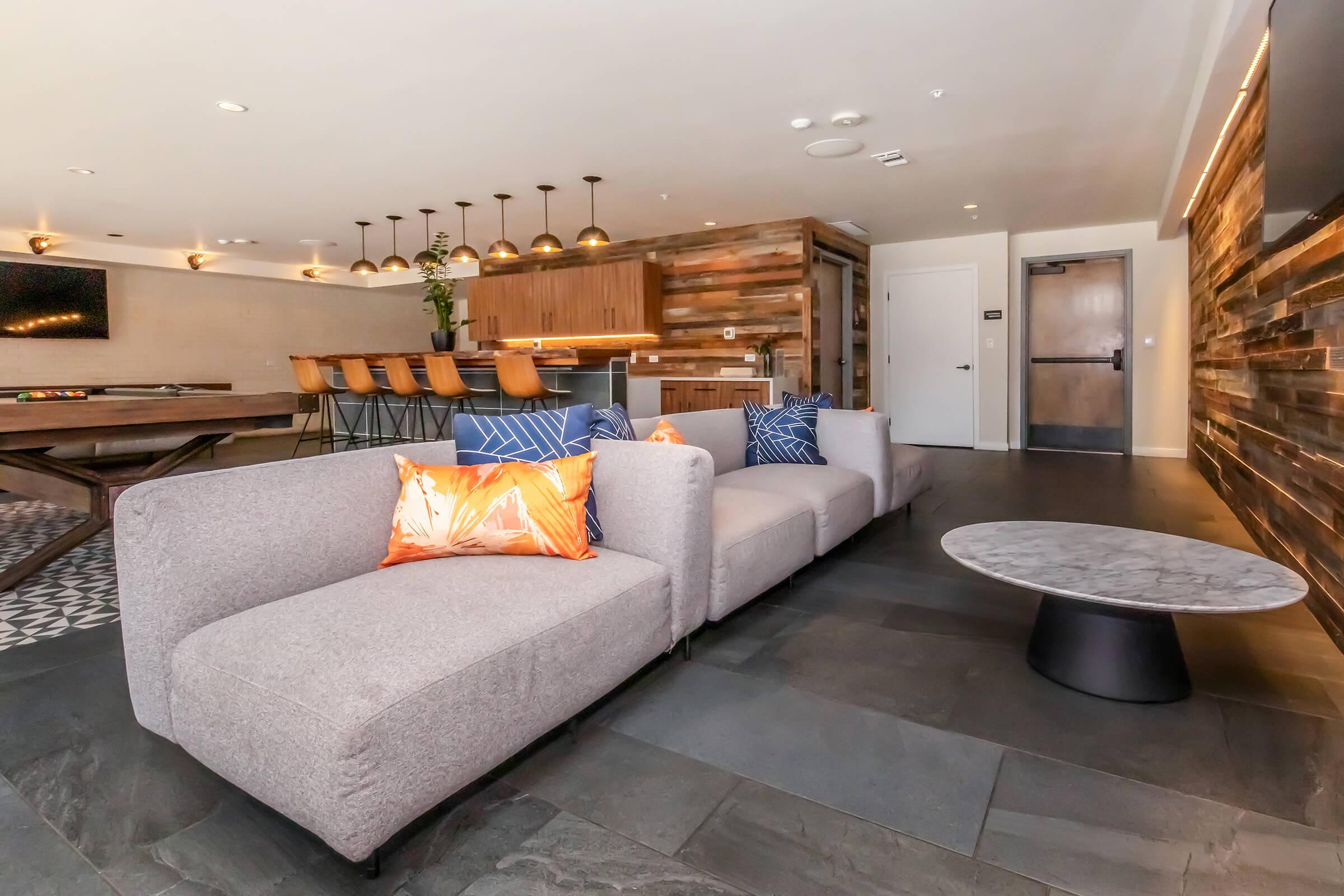 A modern lounge area featuring a gray sectional sofa with colorful throw pillows, a round marble coffee table, and a wooden bar with high stools. The space has a stylish design with wooden paneling on the walls and warm lighting, creating a welcoming atmosphere for relaxation or socializing.