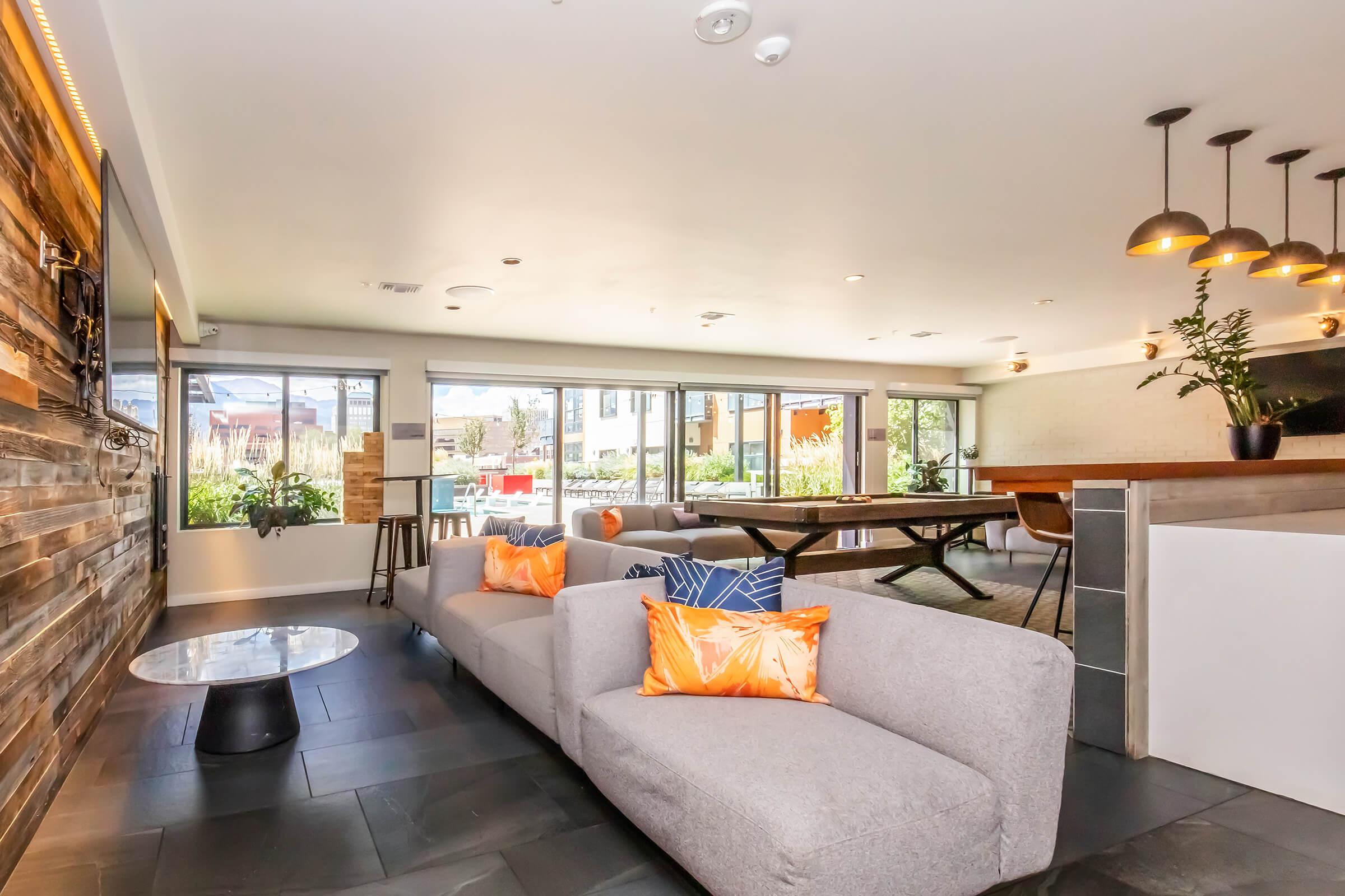 A modern lounge area featuring a gray sofa with orange accent pillows, a round coffee table, and large windows providing natural light. The space includes a dining table, wooden accents on the walls, and a contemporary bar area, creating a cozy and inviting atmosphere for socializing.