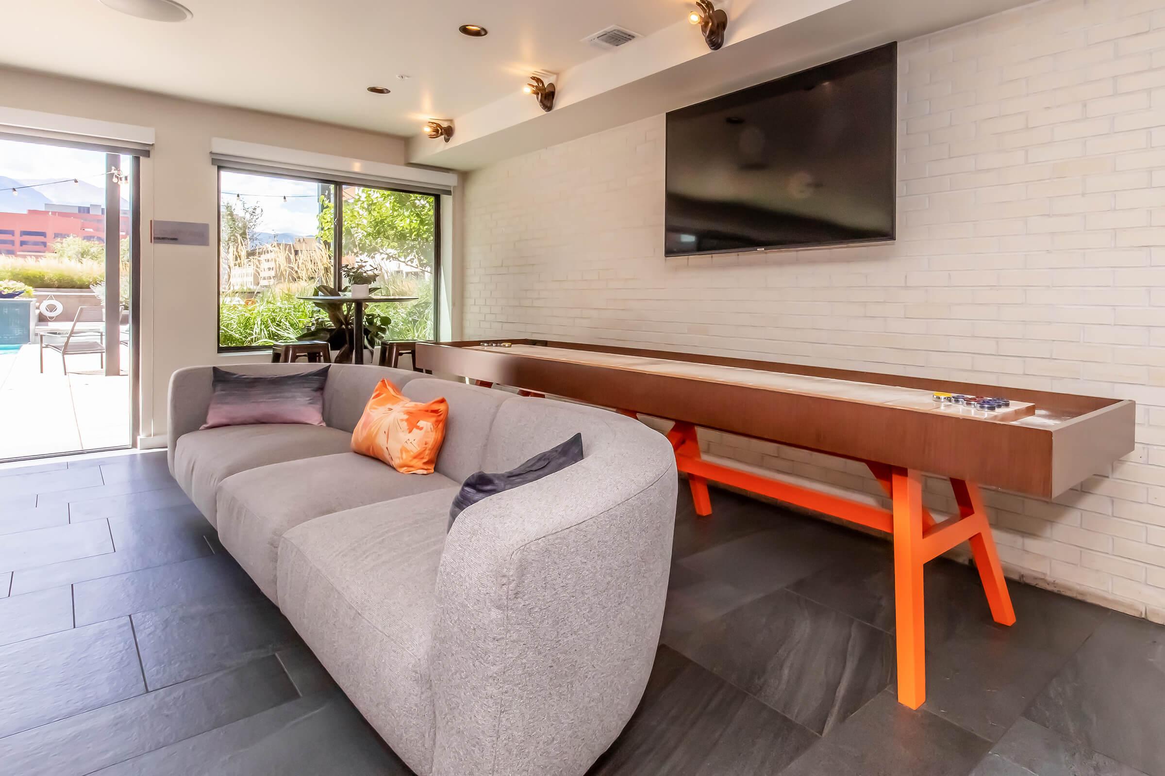 A modern lounge area featuring a gray curved sofa with colorful throw pillows, a stylish orange table, and a large wall-mounted TV. The space includes large windows that offer a view of an outdoor area, adding natural light to the room with a sleek, contemporary design.