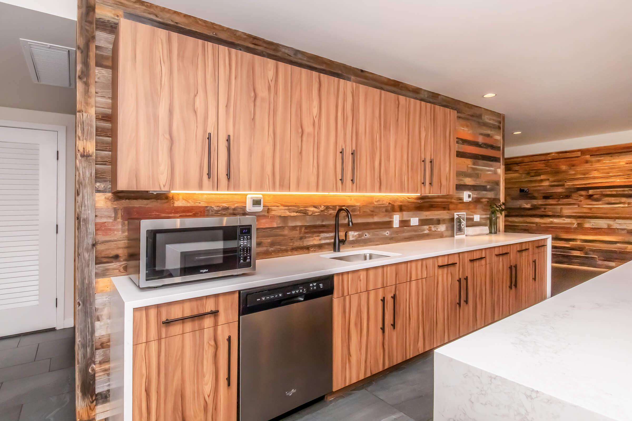 Modern kitchen with wooden cabinetry, a stainless steel dishwasher, microwave, and a white countertop. A sleek sink is installed below a light strip, and a textured wooden wall adds warmth to the design. The space features neutral-toned flooring and stylish, contemporary design elements.