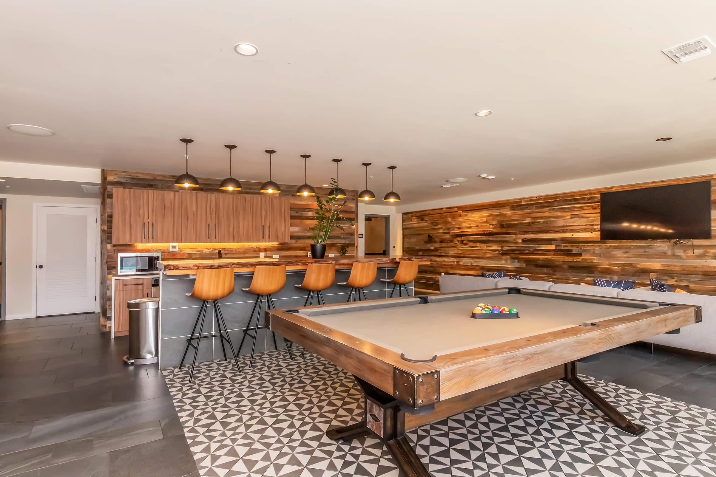 A spacious game room featuring a pool table in the foreground and a bar with high stools in the background. The walls are accentuated with wooden panels, and there is a modern TV mounted on the wall. The floor is a stylish black and white patterned tile. Natural light enhances the inviting atmosphere.