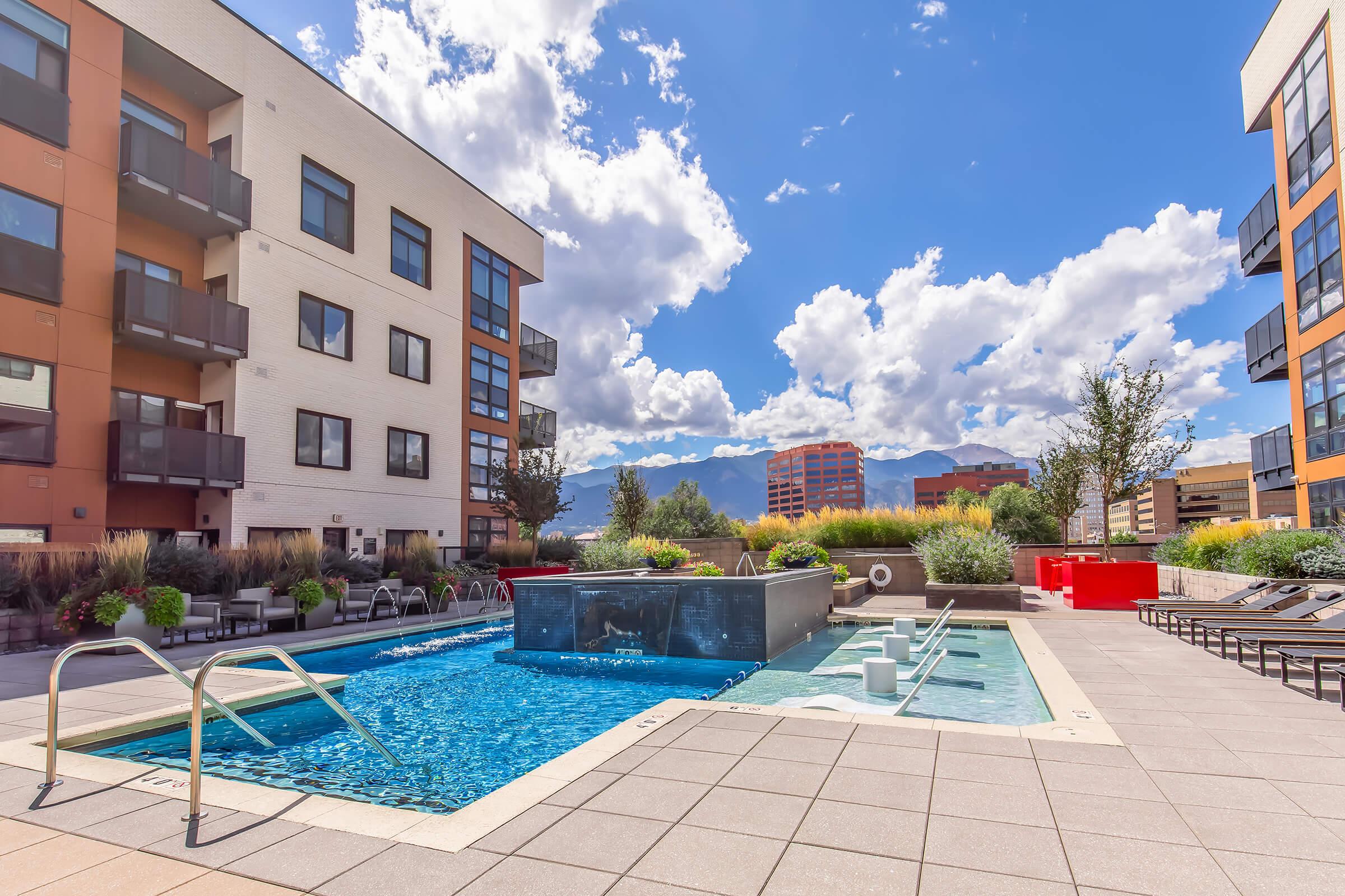 A modern apartment complex featuring a swimming pool surrounded by lounge chairs. The pool area includes a water feature and is set against a backdrop of blue skies with fluffy clouds and distant mountains. The surrounding buildings are sleek and contemporary, with greenery adding to the ambiance.