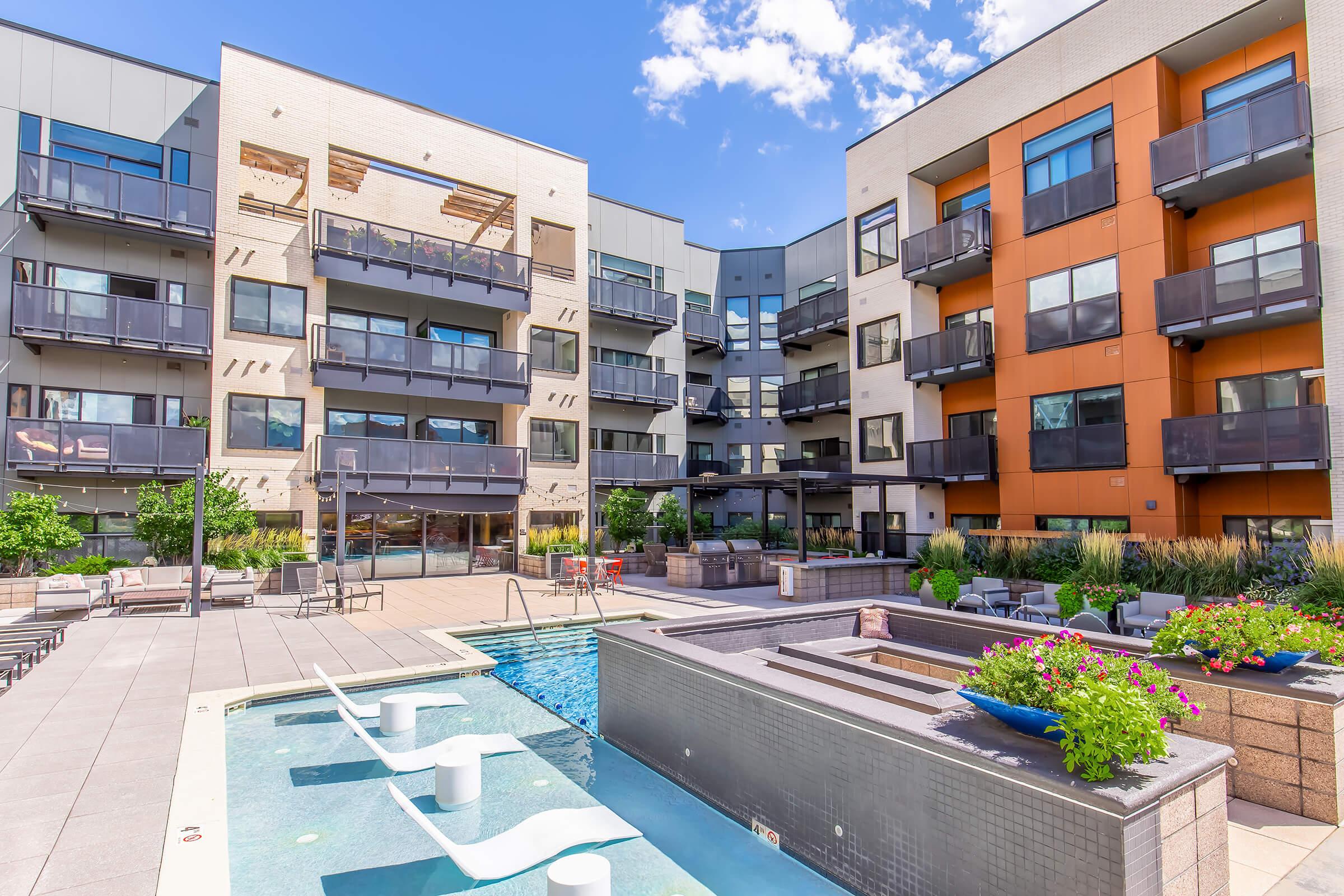 Modern apartment complex with a sleek design, featuring multiple balconies, a swimming pool, and landscaped outdoor spaces. Bright blue sky and scattered clouds enhance the inviting atmosphere, making it an appealing residential area for potential residents.