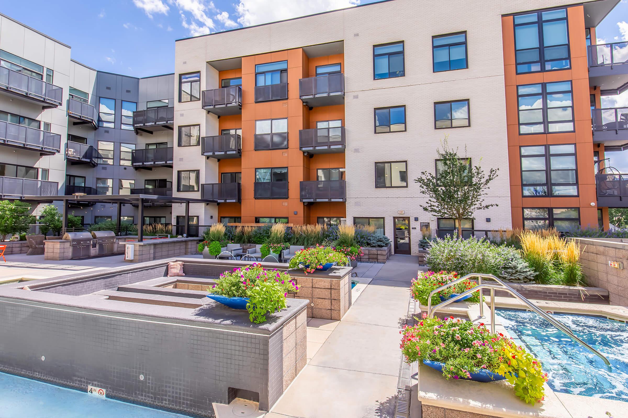 Modern apartment complex featuring a landscaped courtyard with colorful flower beds, a swimming pool, and a hot tub. The building has multiple levels with balconies and a mix of brick and contemporary architecture. Clear blue sky and greenery enhance the inviting atmosphere.