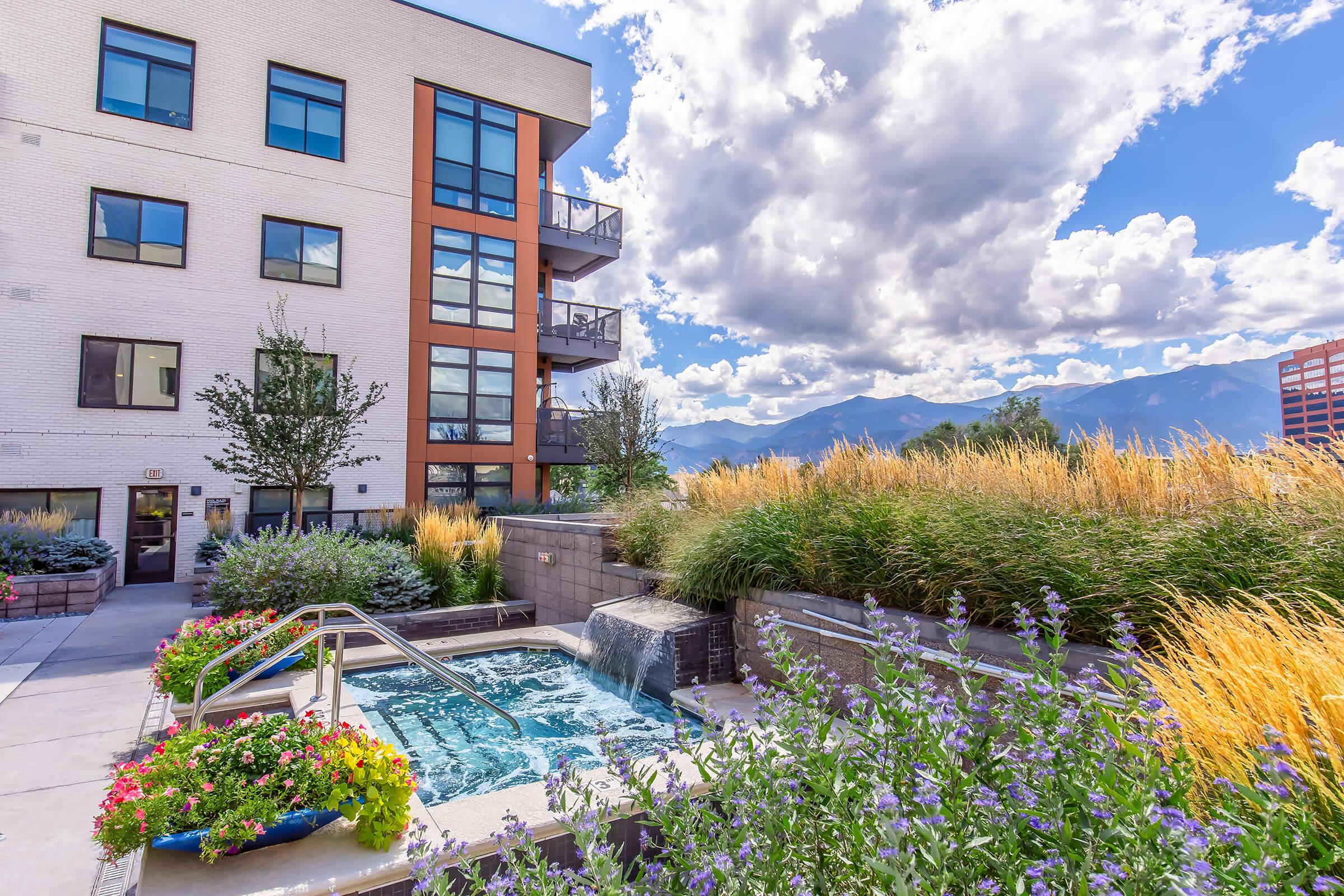 A tranquil outdoor area featuring a small hot tub surrounded by colorful flowers and greenery, with a waterfall. In the background, a modern building with balconies and scenic mountains under a partly cloudy sky. Ideal setting for relaxation and enjoyment of nature.