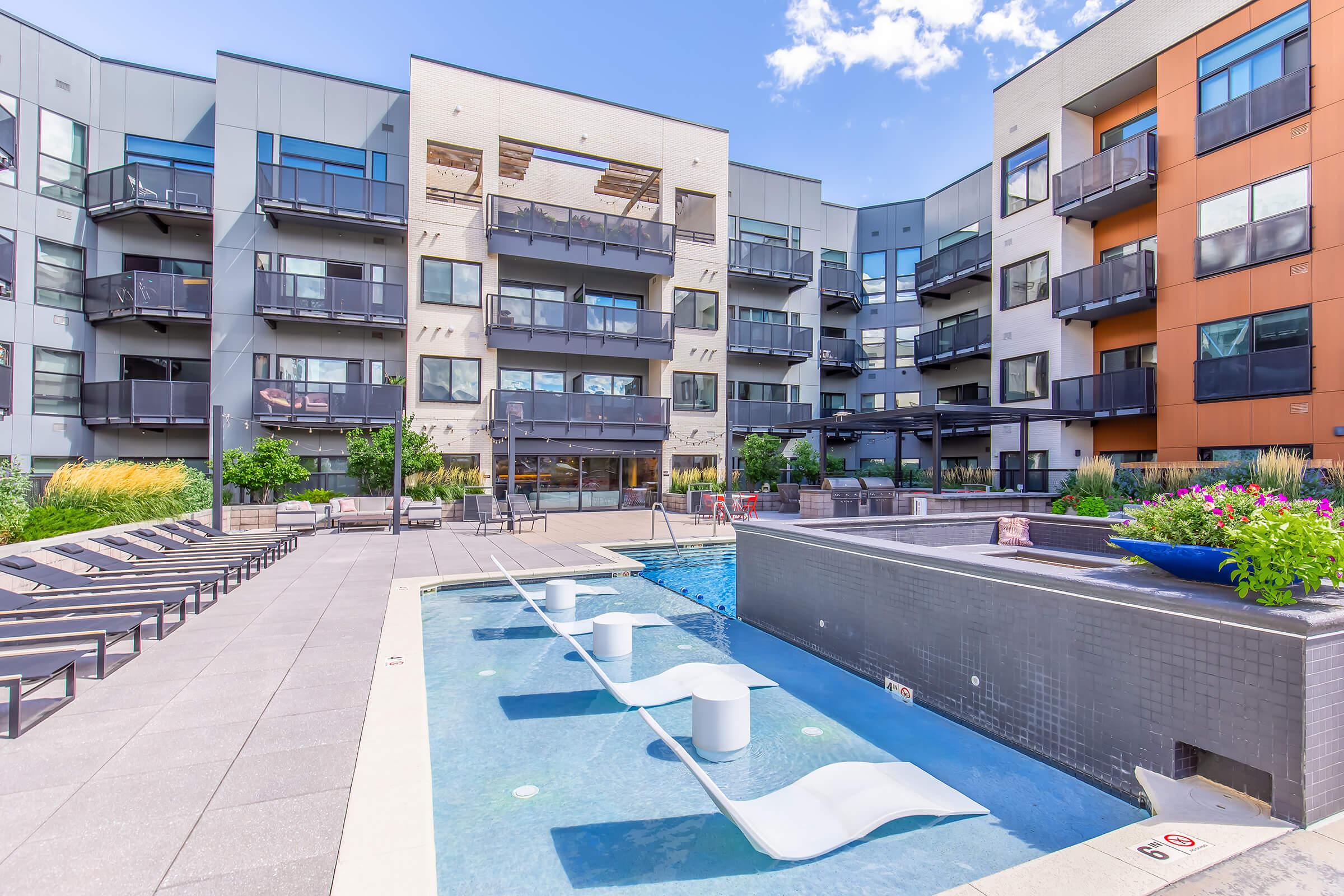 A modern apartment complex featuring a swimming pool with lounge chairs, landscaped areas, and balconies. The building has a mix of neutral and warm-colored facades, and there are potted plants around the pool area, with a clear blue sky overhead.